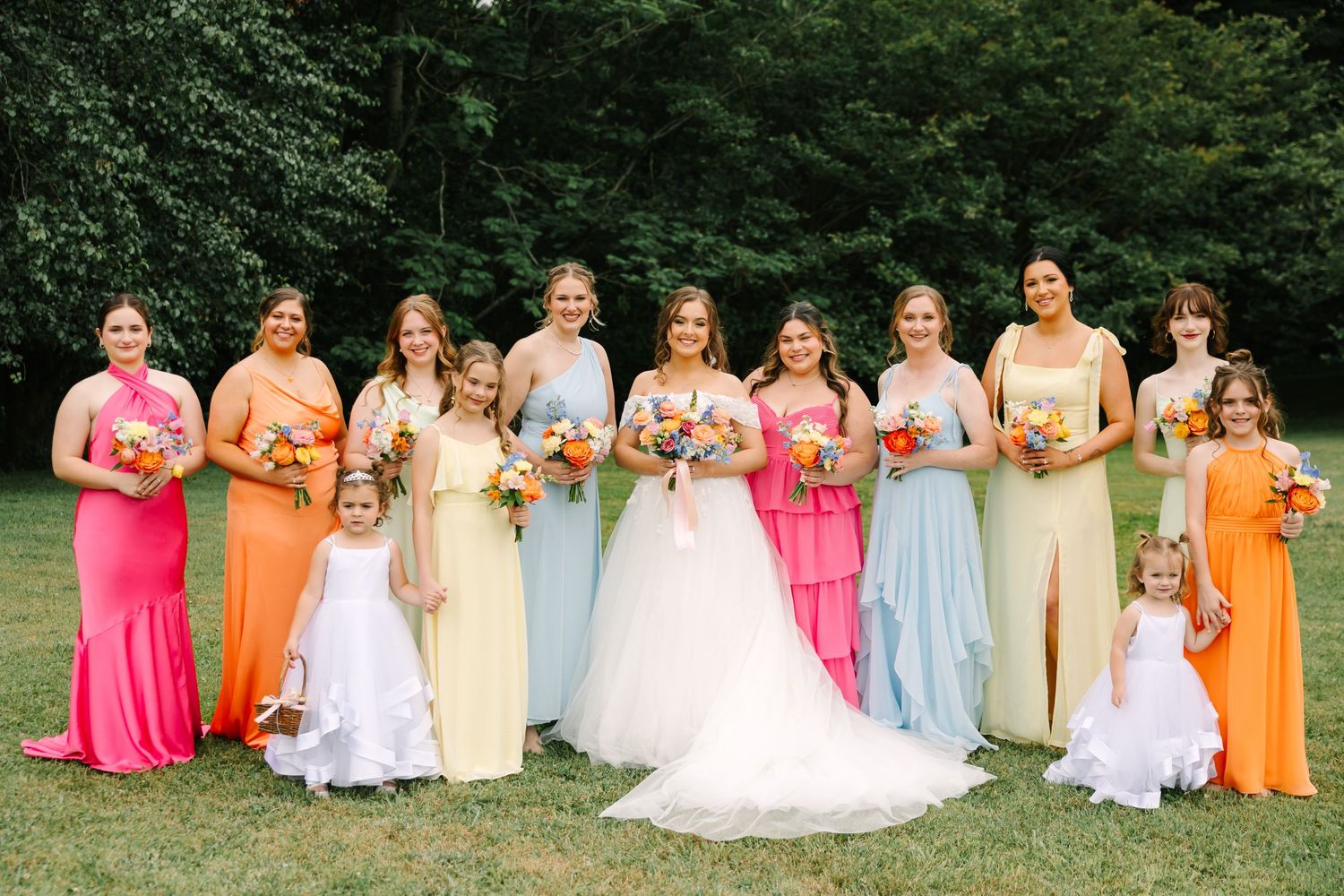 Bridesmaids in colorful pink, orange, and light blue dresses pose with the bride on a grassy lawn during summer.