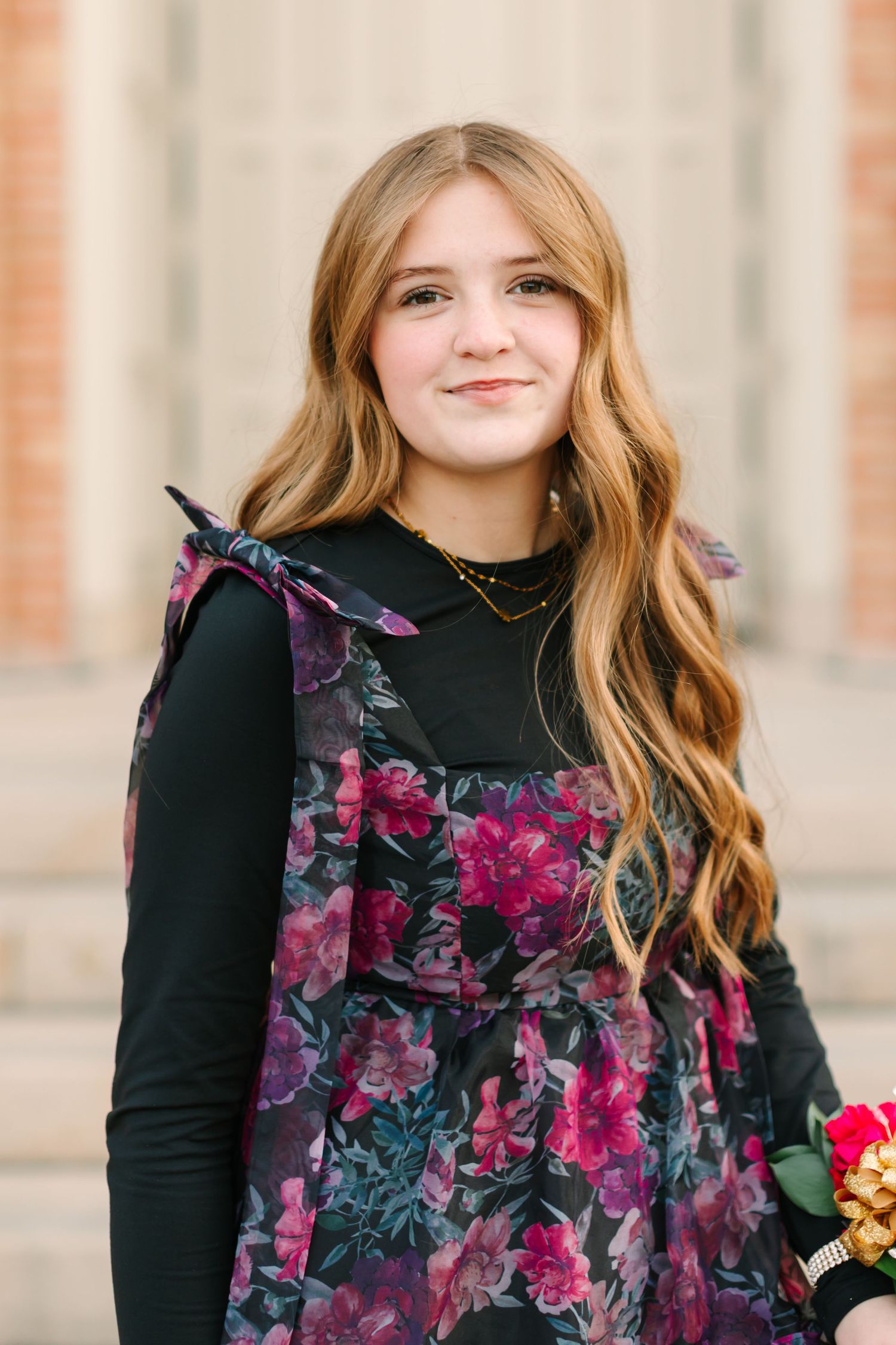 A person in a black turtleneck and floral dress poses against a brick wall backdrop.