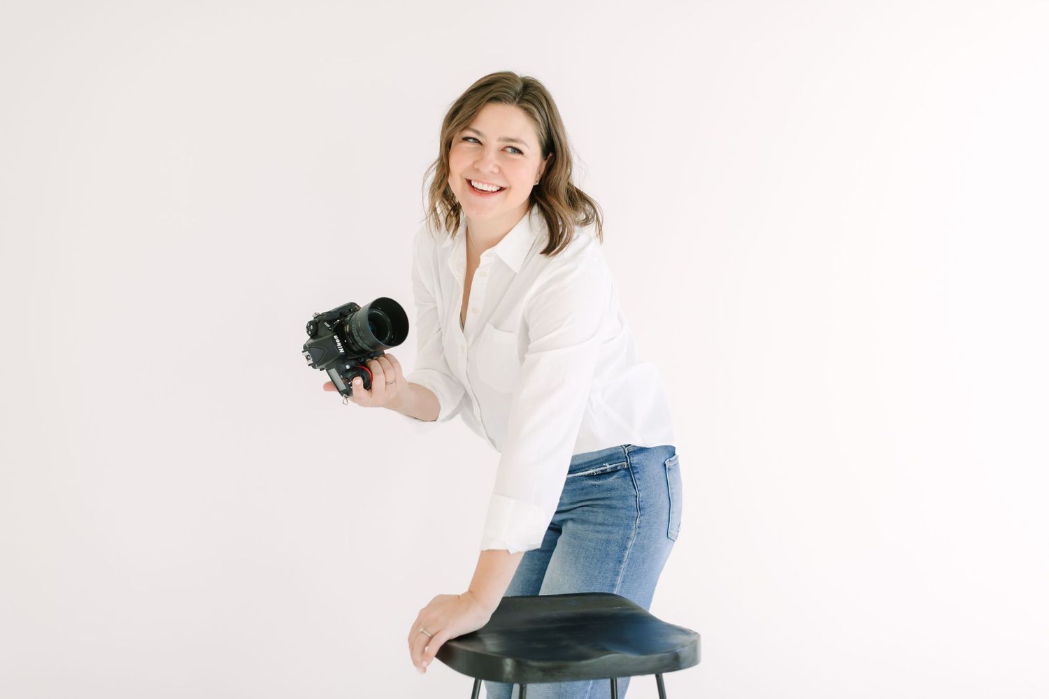 Professional photographer in white shirt and jeans kneels on stool while holding DSLR camera against white background.