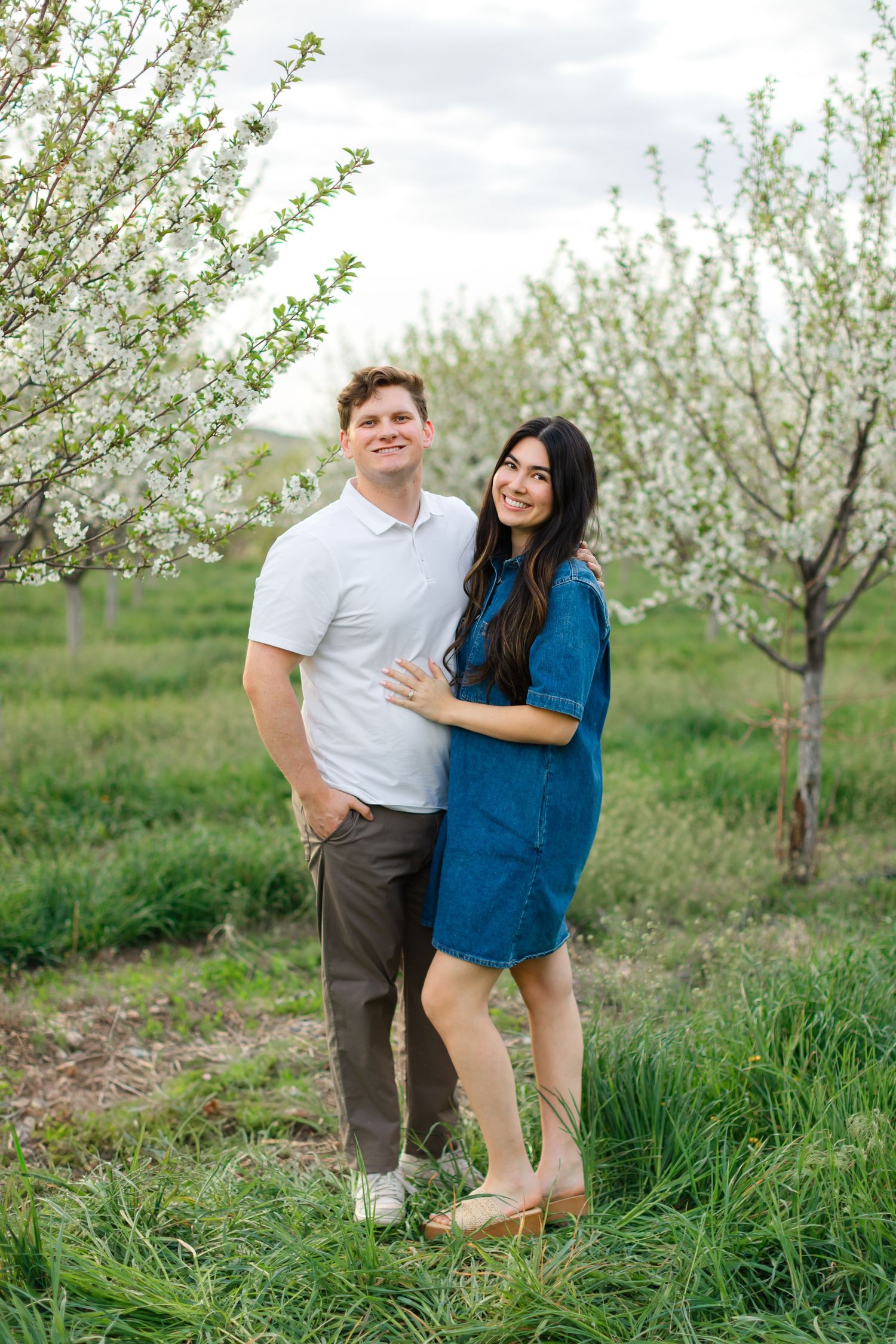 A couple poses together in a blooming spring orchard with white blossoms and green grass.
