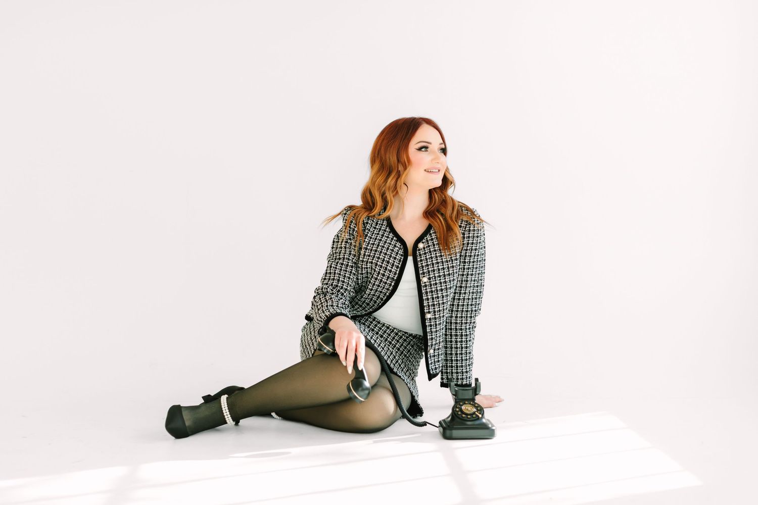 Model in plaid shirt and tights sits casually on white backdrop while looking to the side.