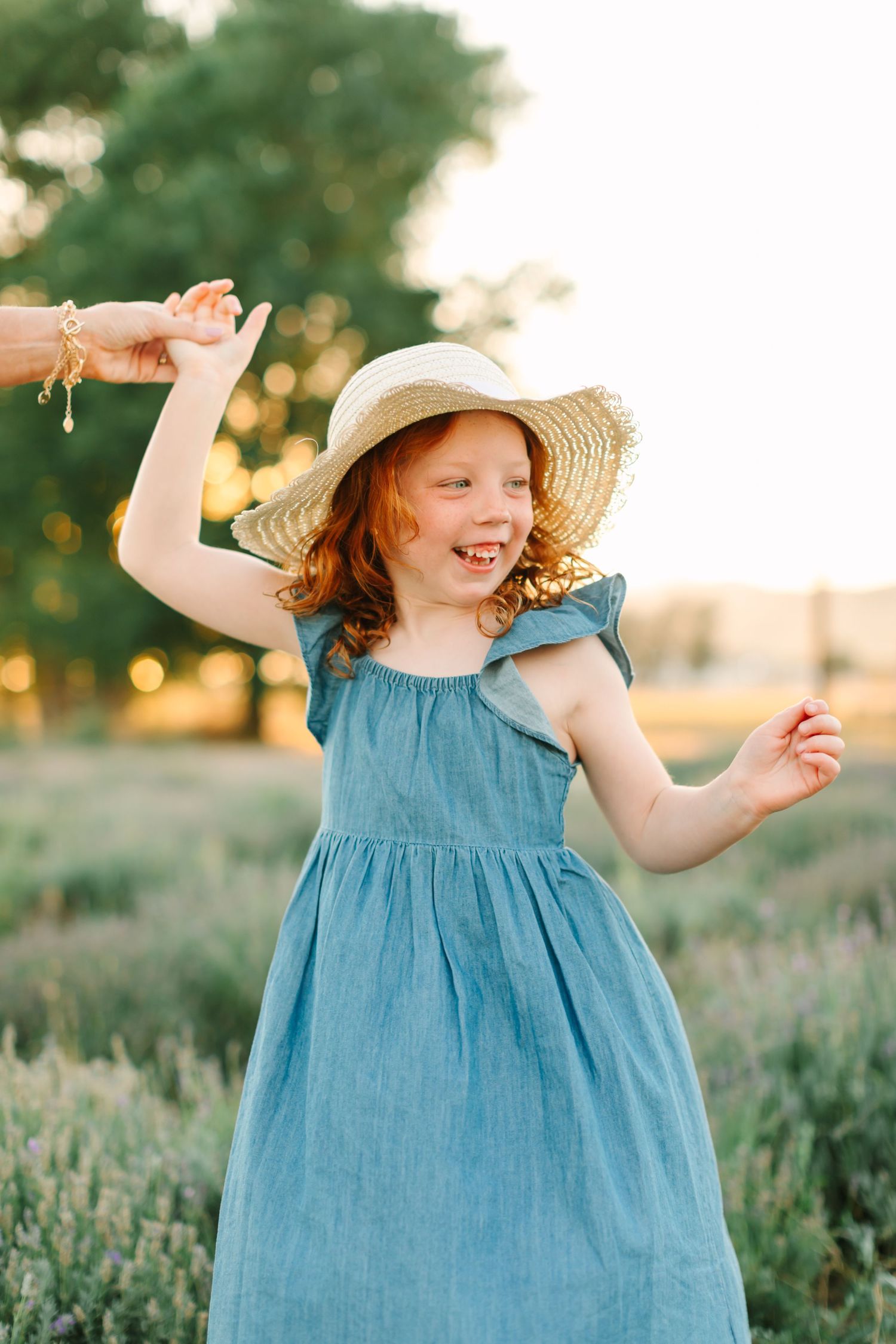 Child in blue dress and straw hat playing joyfully in a lavender field at sunset.