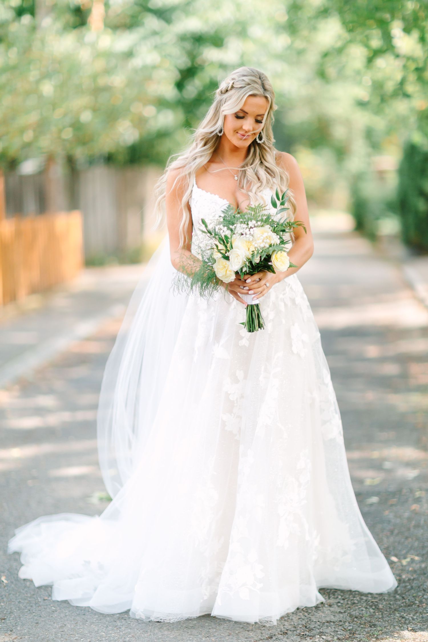 A bride in a flowing white wedding dress holds a bouquet of white flowers in a sunlit outdoor garden setting.