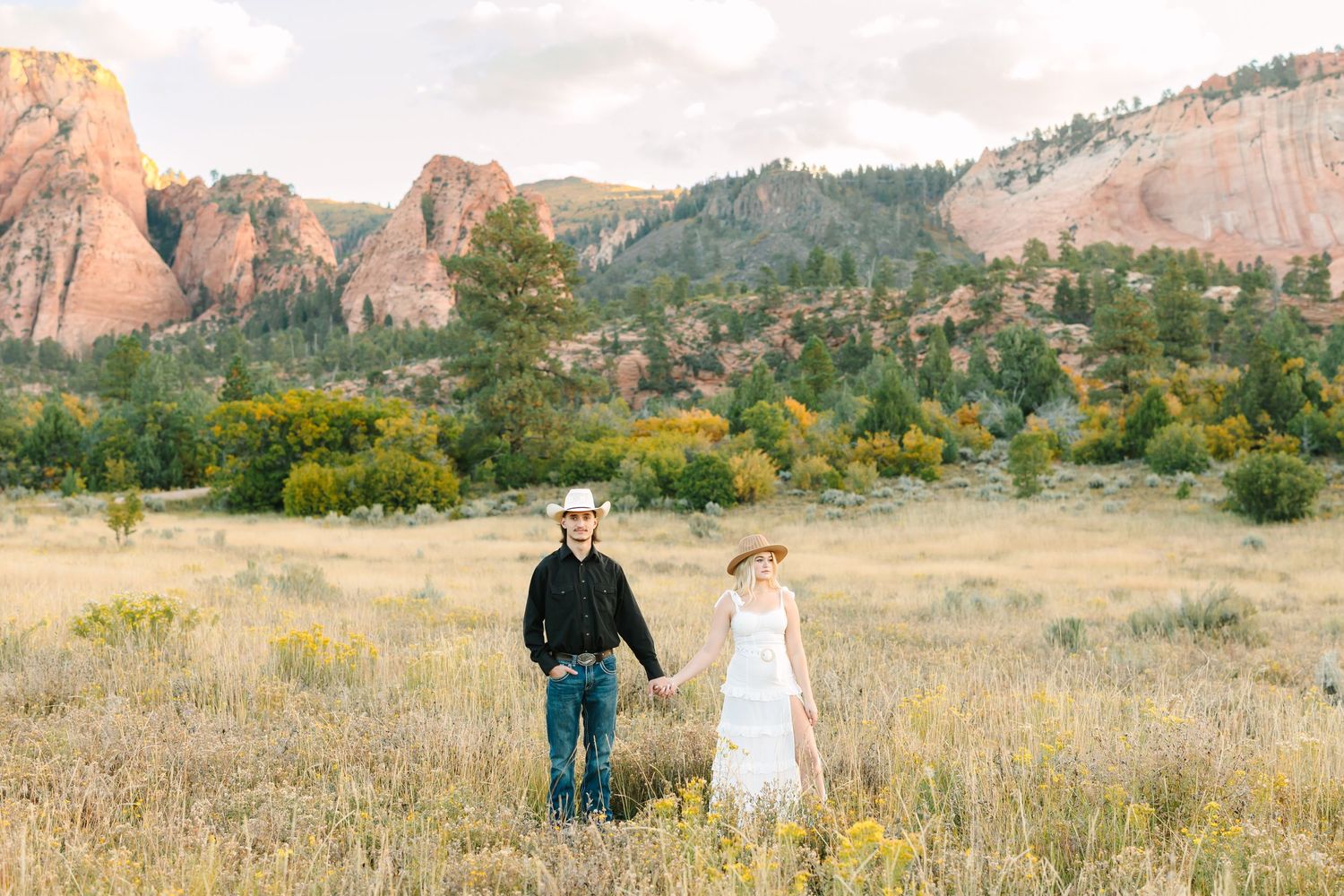 Couple walking hand in hand through a scenic meadow with towering red rock formations and mountains in the background.