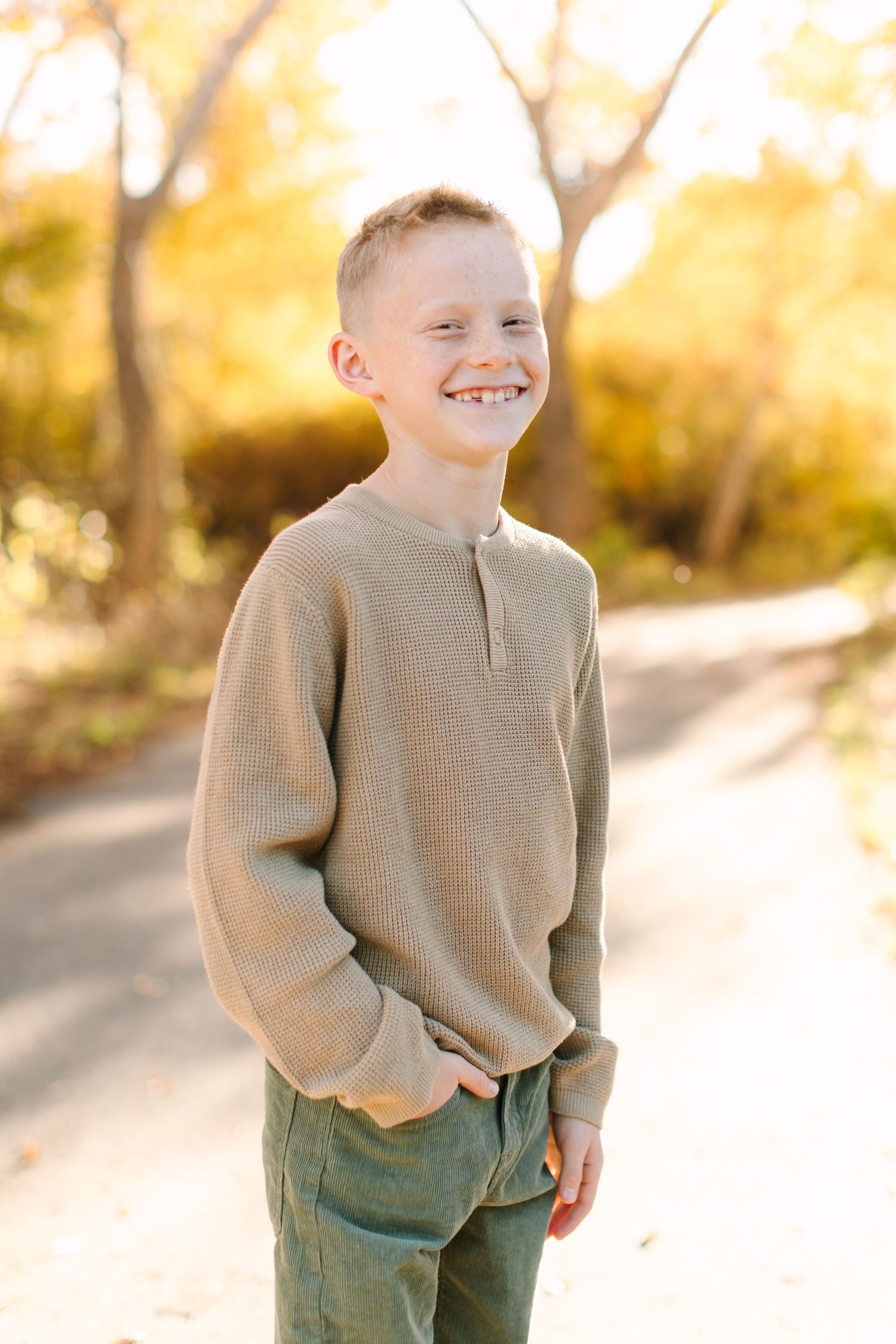 A child in a beige sweater and green pants stands on a sunlit path during autumn.