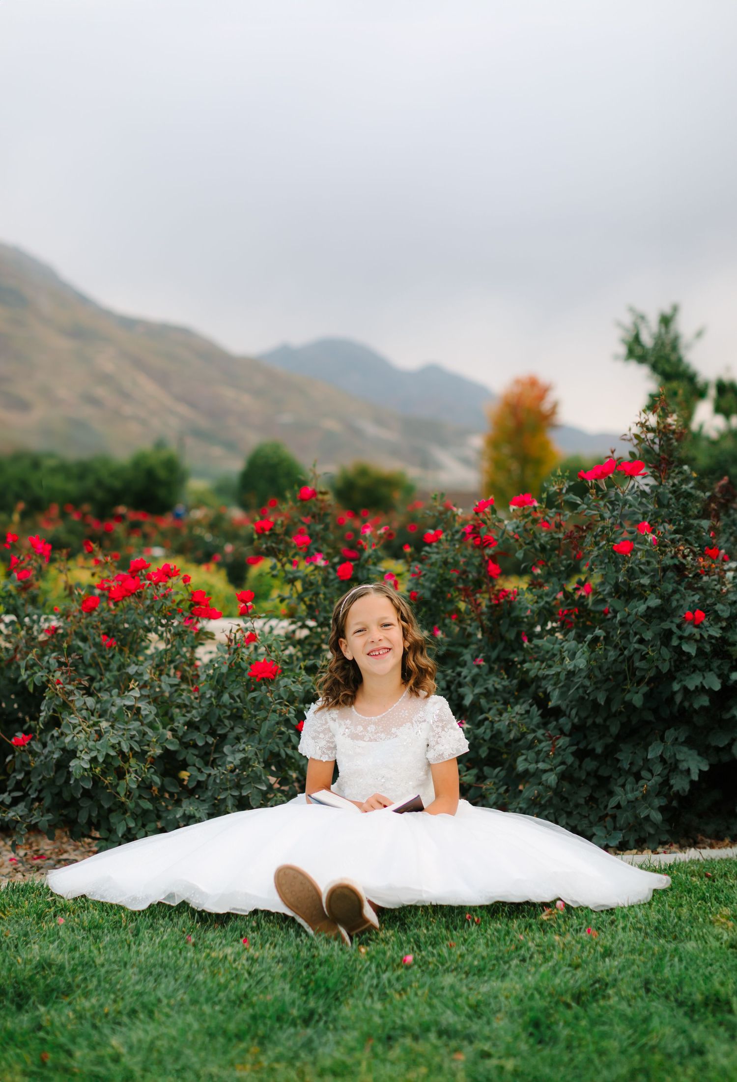 A white dress spreads across green grass with red roses in the background against mountain scenery.