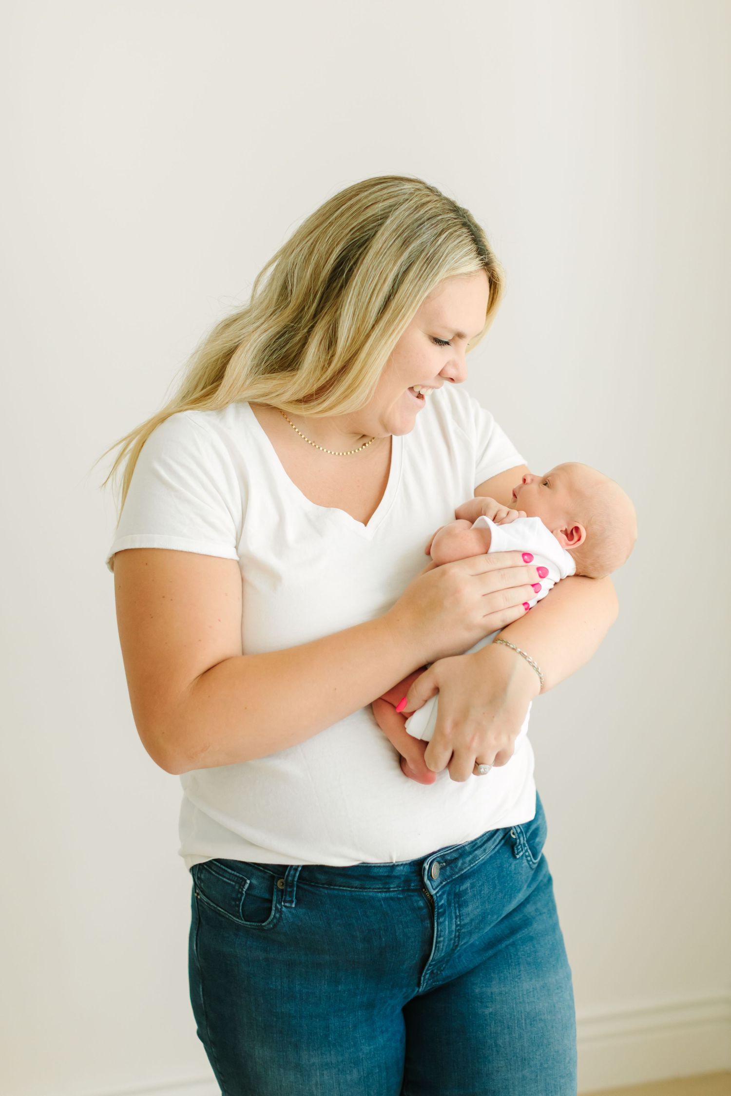 Mother in white shirt and jeans holds and snuggles with newborn baby against light background.