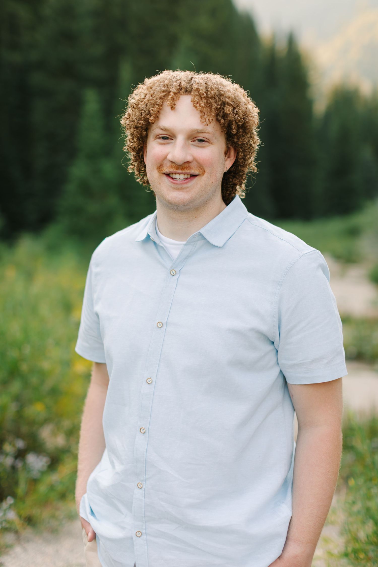 Smiling young man with curly red hair and mustache wearing a light blue shirt outdoors in a mountain meadow in Utah.