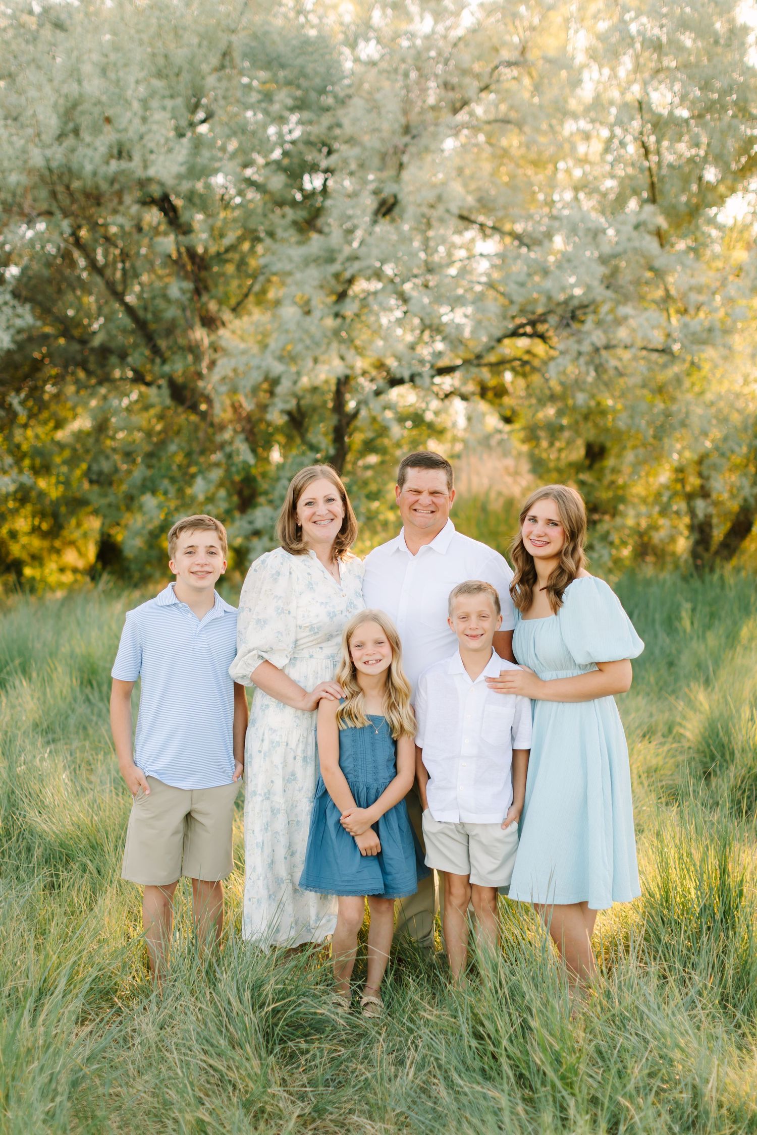 Family of six posing outdoors in a green meadow, wearing coordinated light blue and white outfits, smiling together.