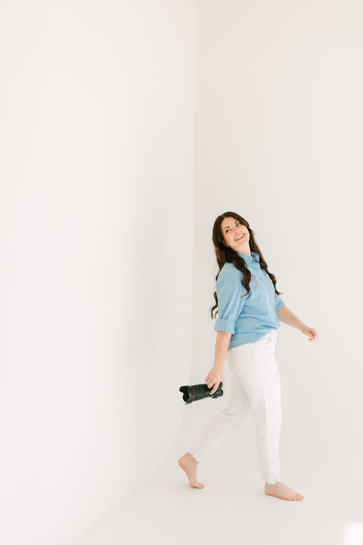 Photographer in casual attire poses playfully while holding camera in minimalist white studio.