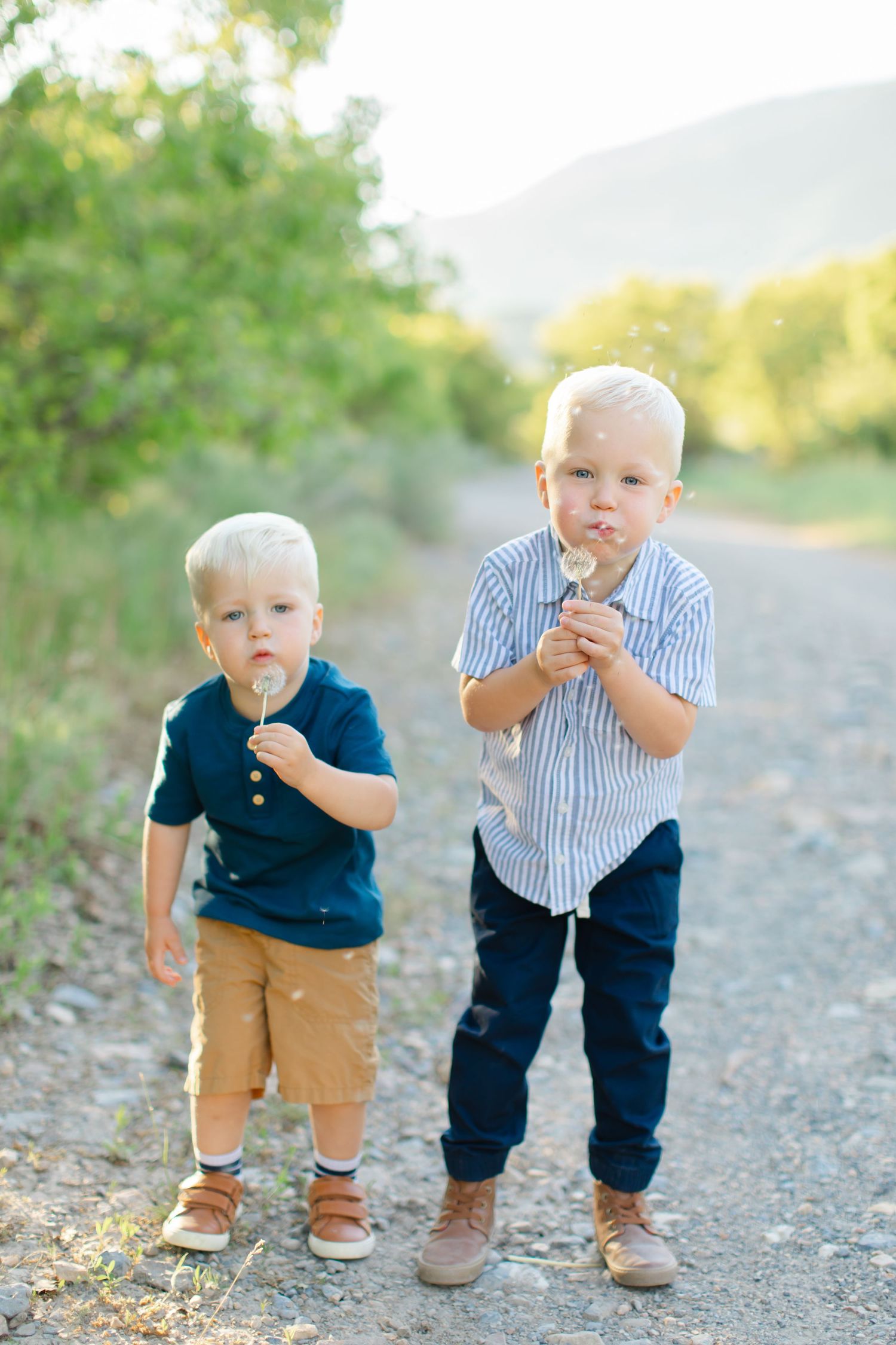 Two young children in casual clothes stand on a dirt path while enjoying lollipops on a sunny day.