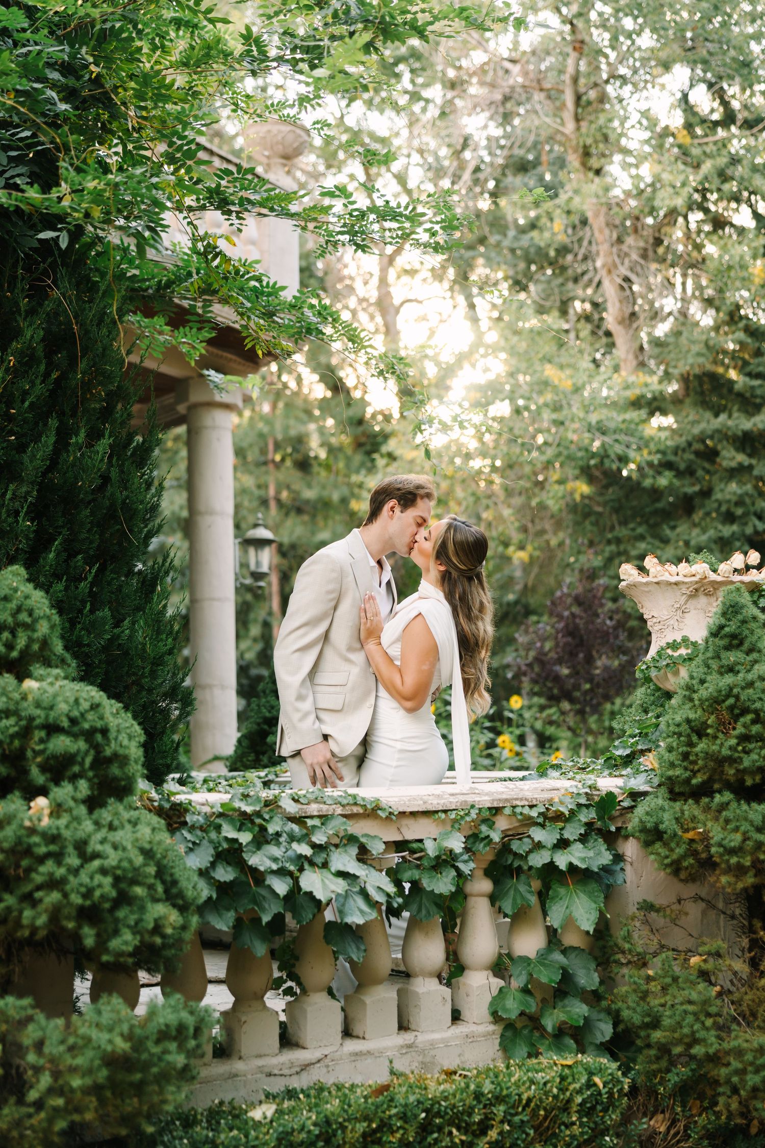 A couple shares a romantic moment on an ivy-covered stone balcony in a lush garden setting at sunset.