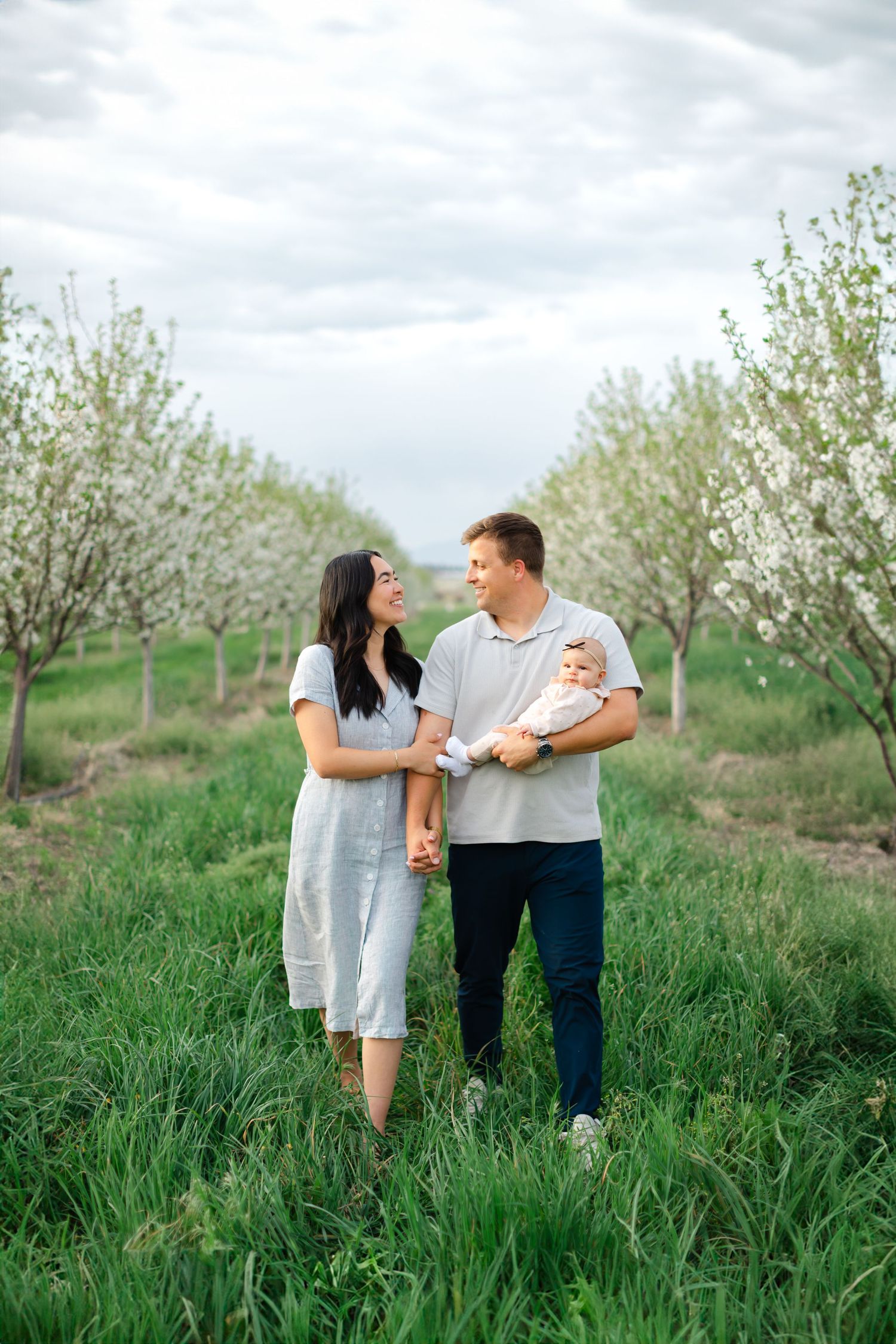 A couple shares a tender moment while walking through a blooming spring orchard on a cloudy day.