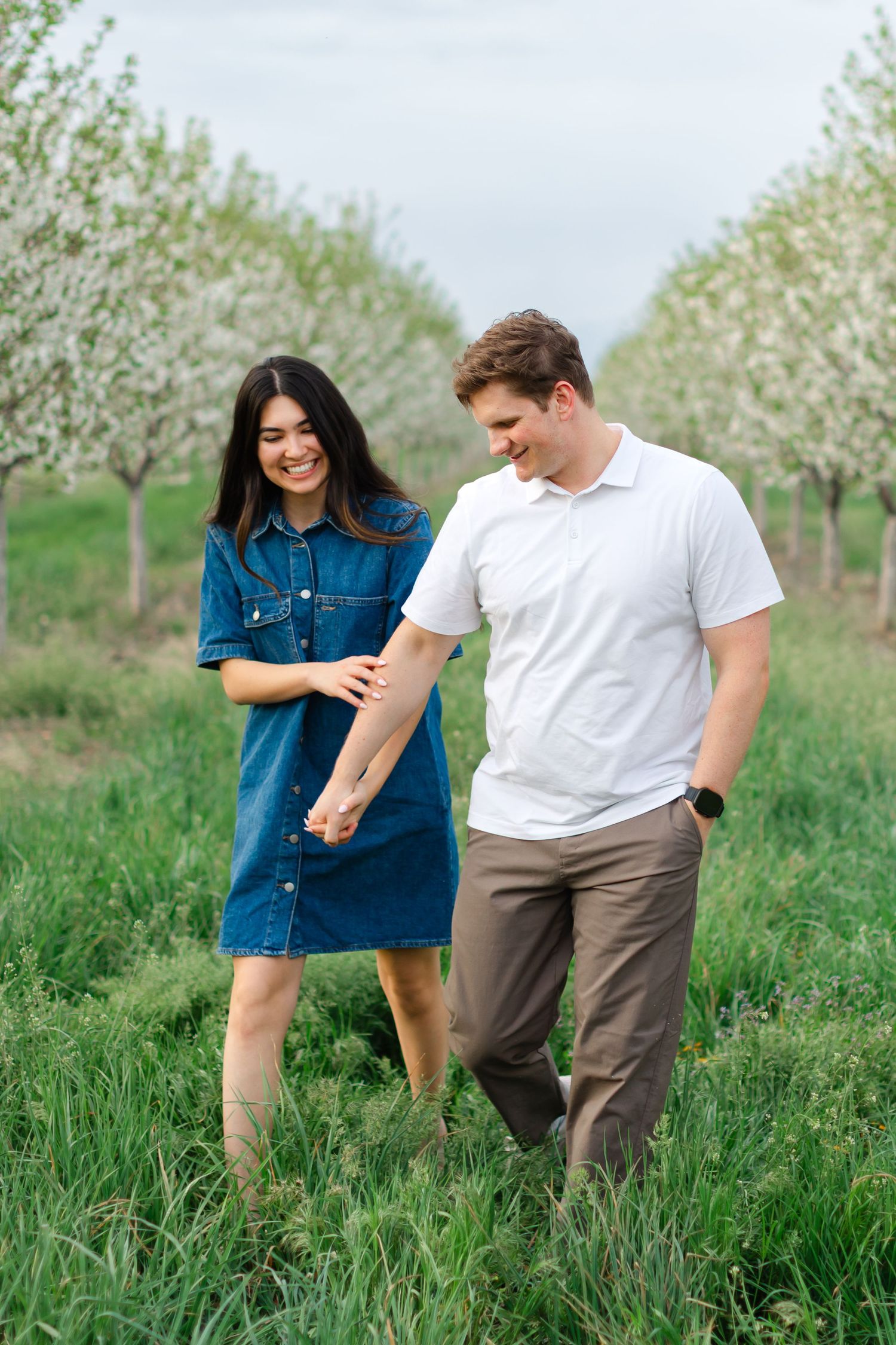 A couple shares a tender moment while walking through a blooming spring orchard on a cloudy day.