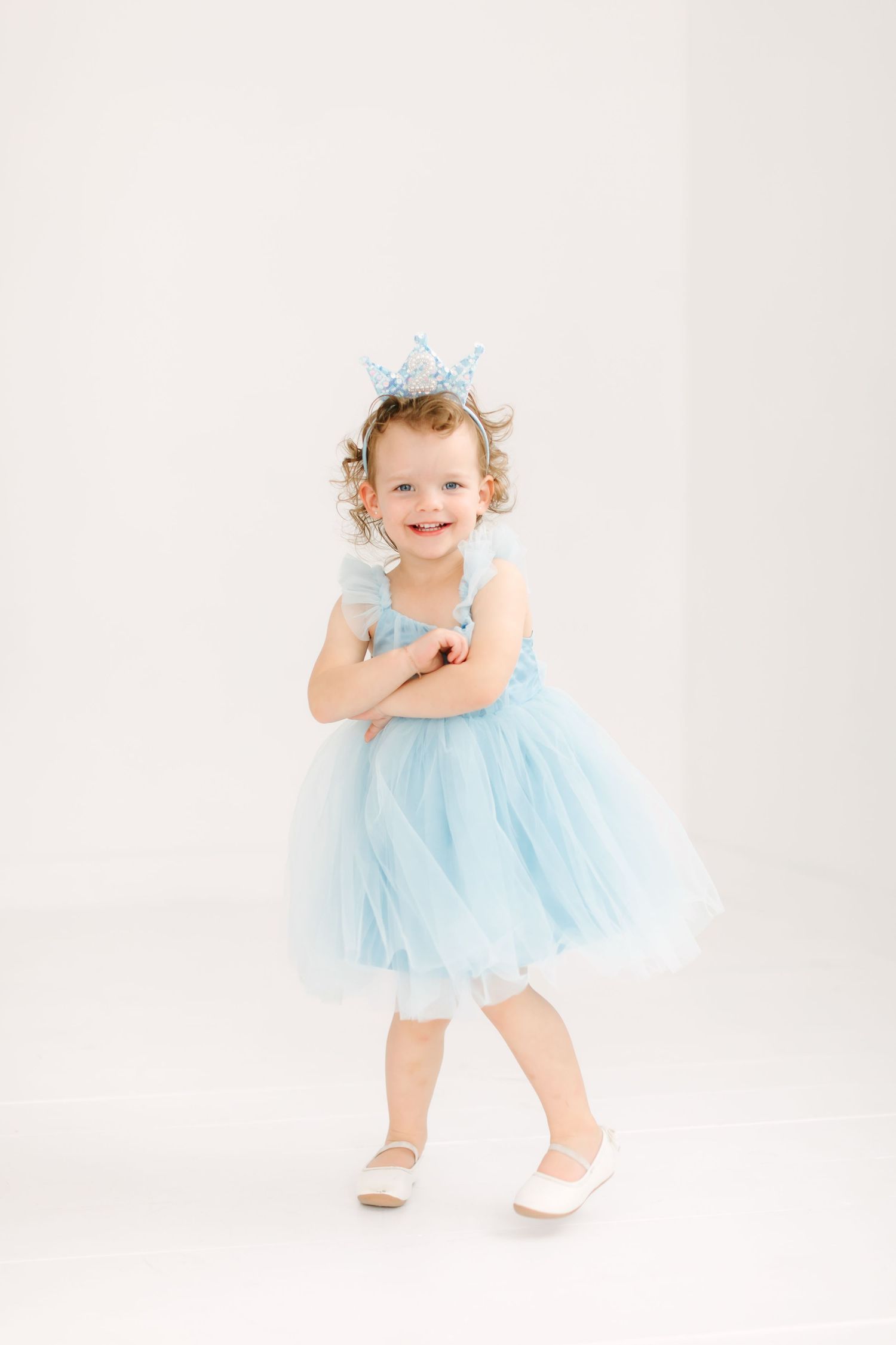 A young girl wearing a blue tulle dress and a crown-like headpiece smiles and stands with her arms crossed.