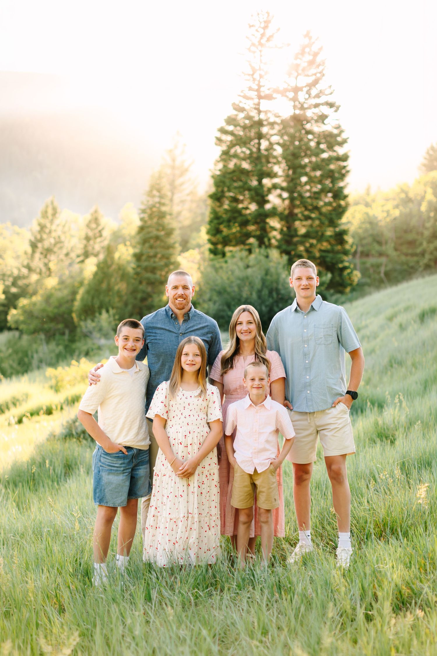 Family of six poses in a sunlit green meadow with tall pine trees, wearing coordinated pastel outfits for a portrait.