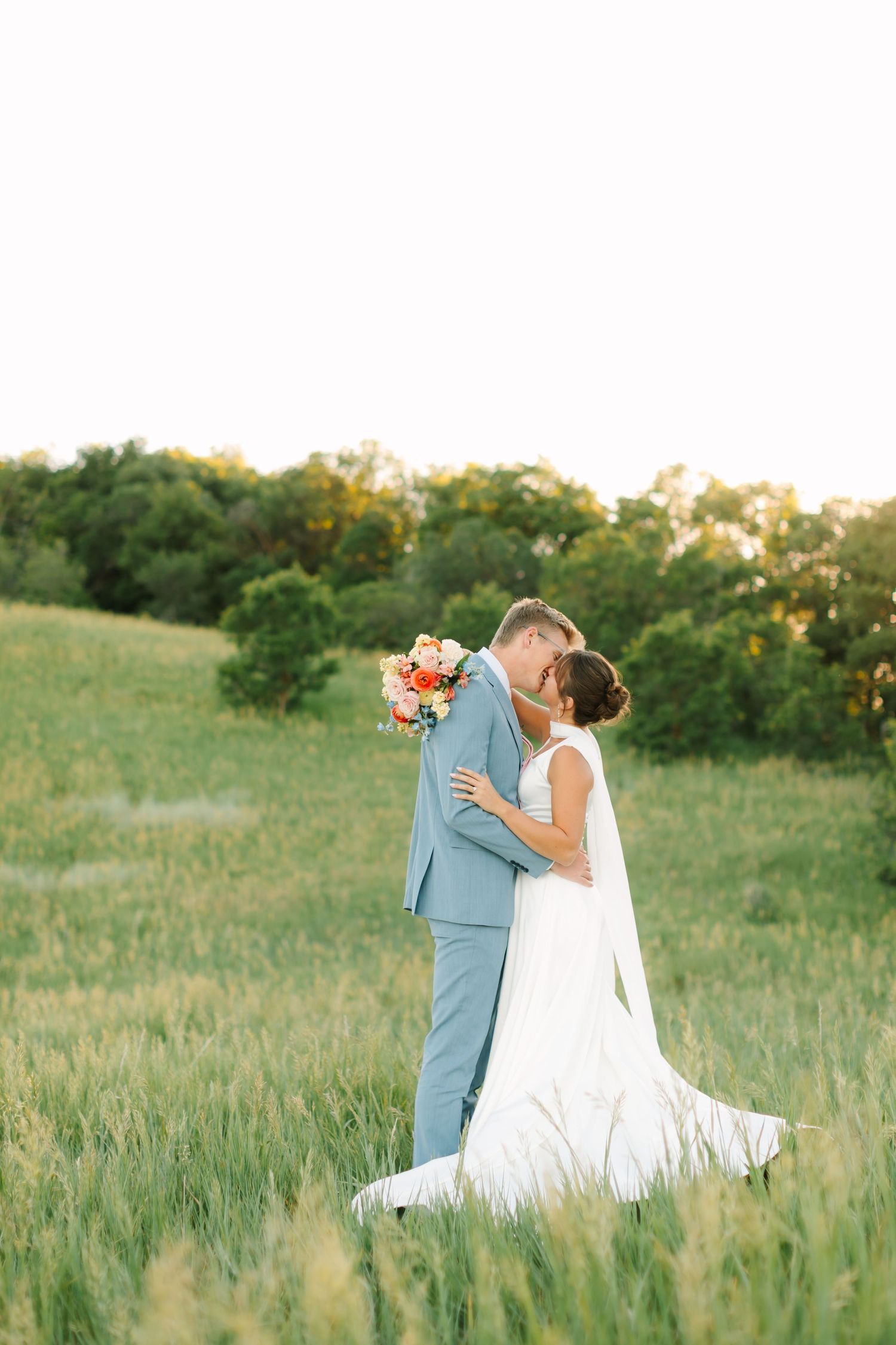 Bride and groom kissing in a green field, bride in white gown with veil holding colorful bouquet, groom in light blue suit.