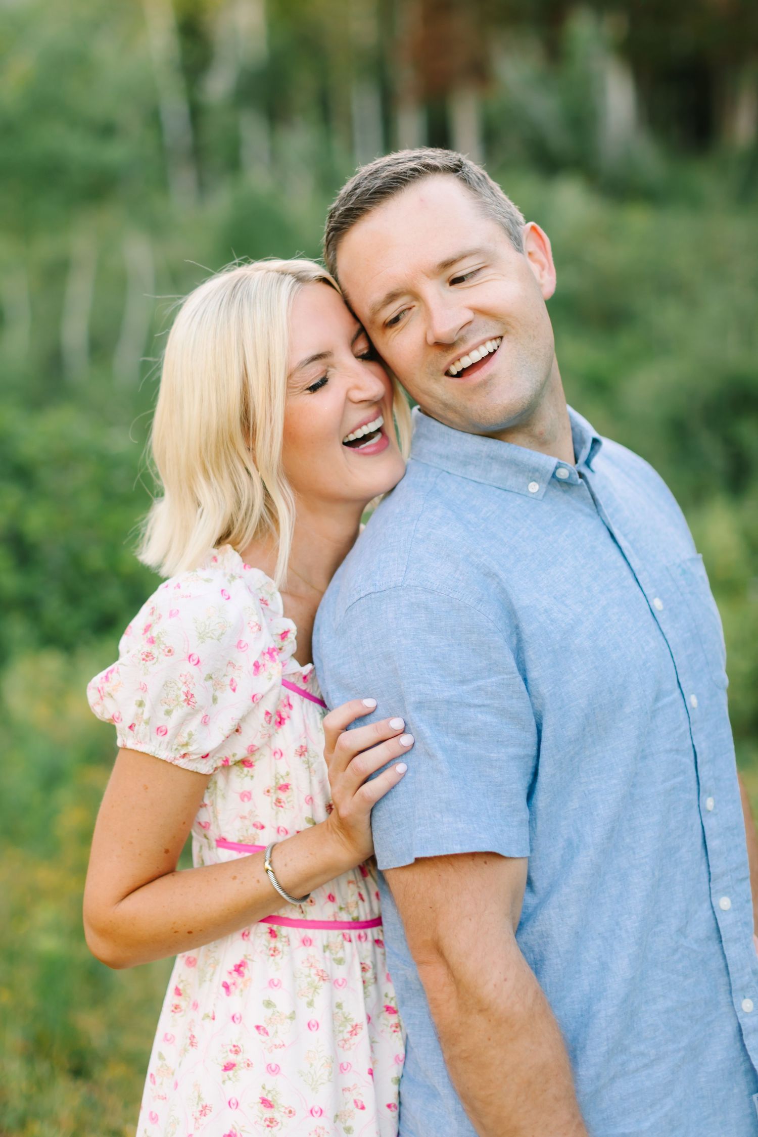 A smiling couple embraces outdoors, woman in floral dress leaning on man in blue shirt, with lush green trees behind.