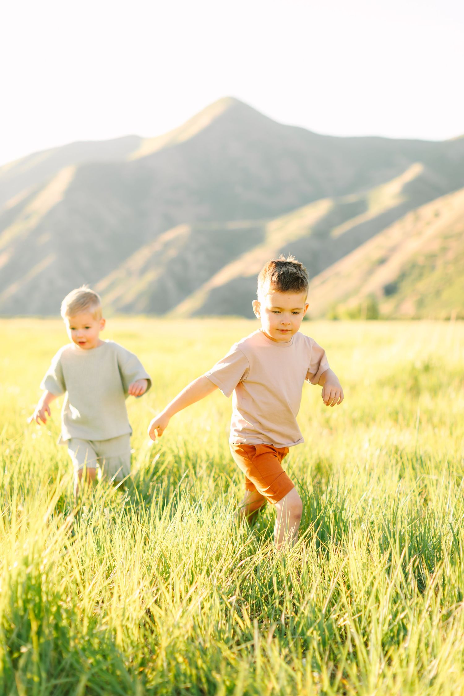 Two young children running through tall green grass with sunlit mountains in the background on a sunny day.