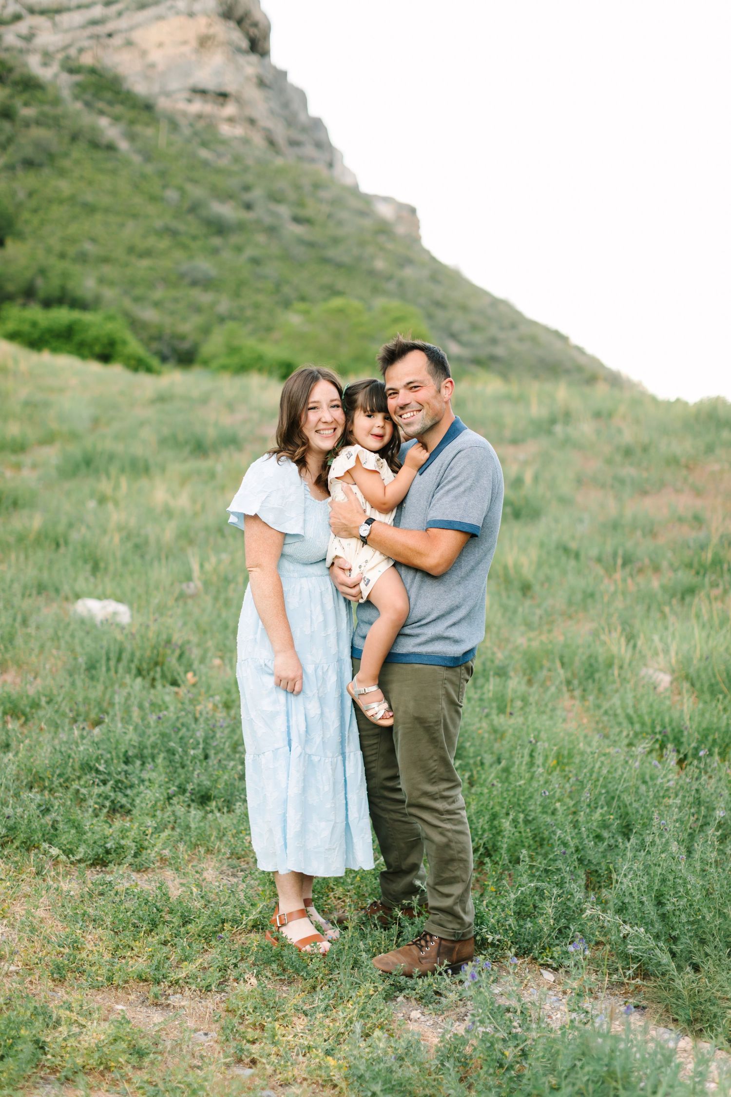 A happy family of three poses outdoors in a green meadow with mountain backdrop, parents holding young child.