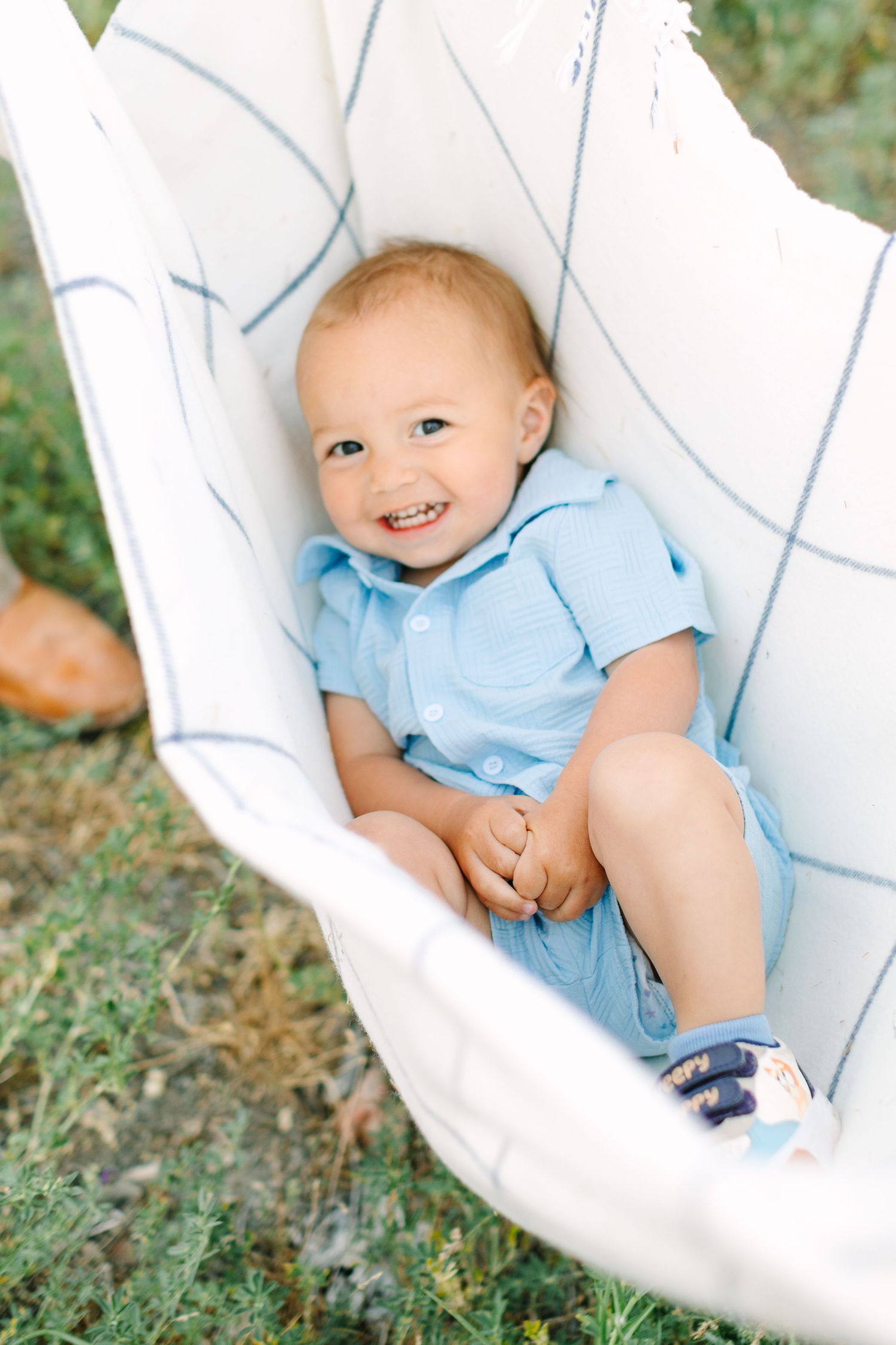 Smiling baby boy in blue romper sitting happily in a white hammock outdoors on a sunny day.