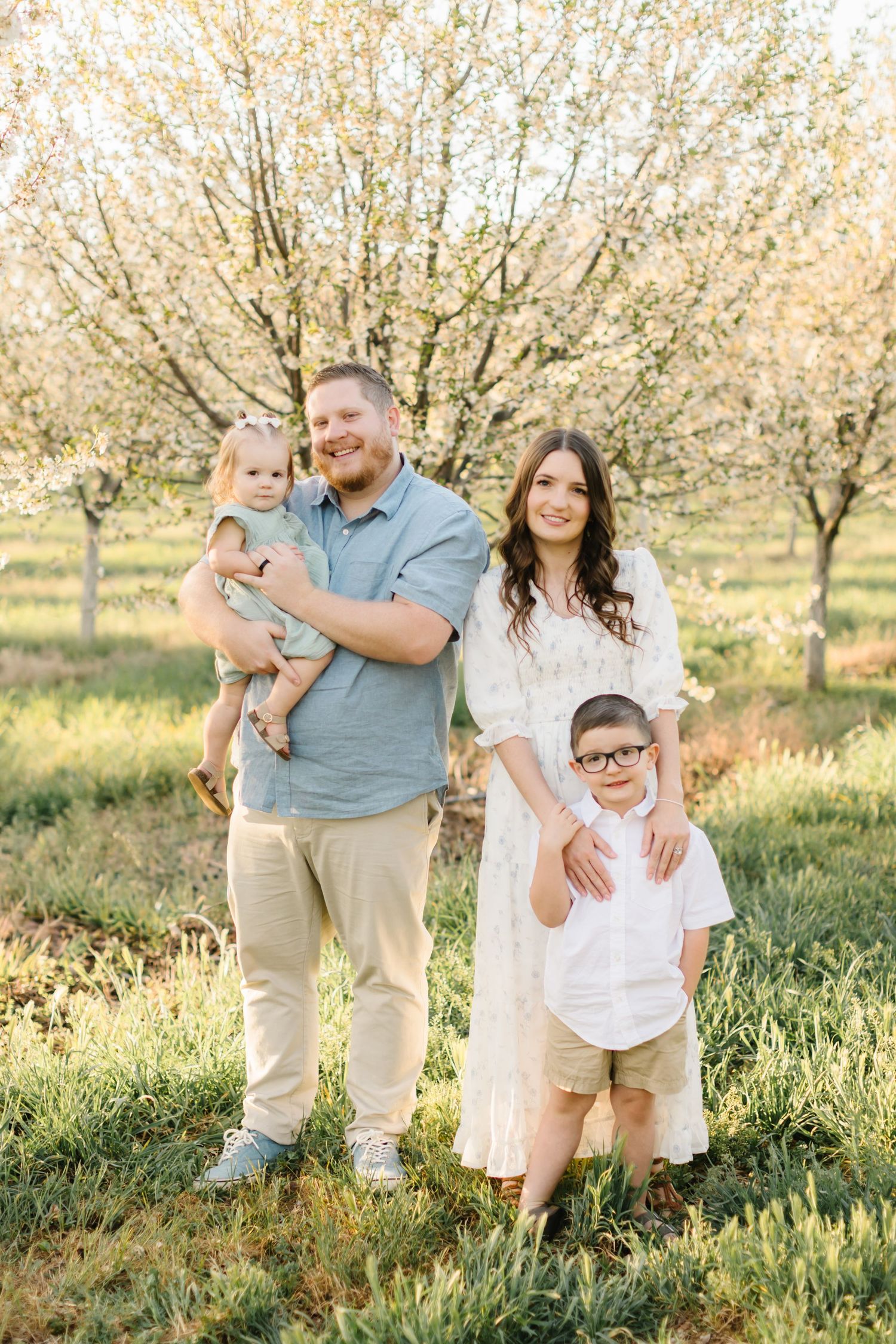 Family of four posing outdoors among blooming spring trees, parents with toddler and young boy in glasses.