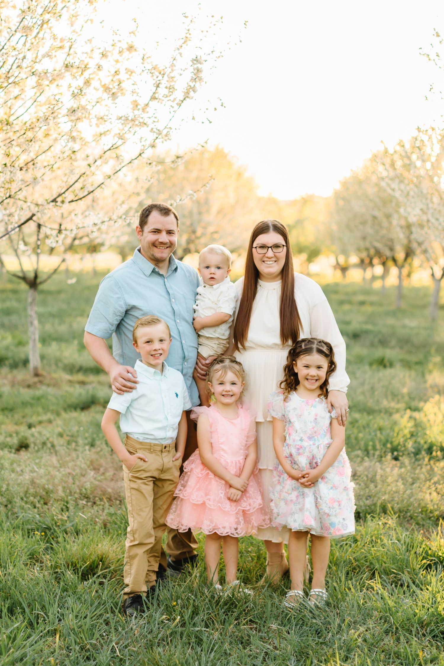 Family of six poses in blooming orchard at golden hour, parents with four young children in spring attire.