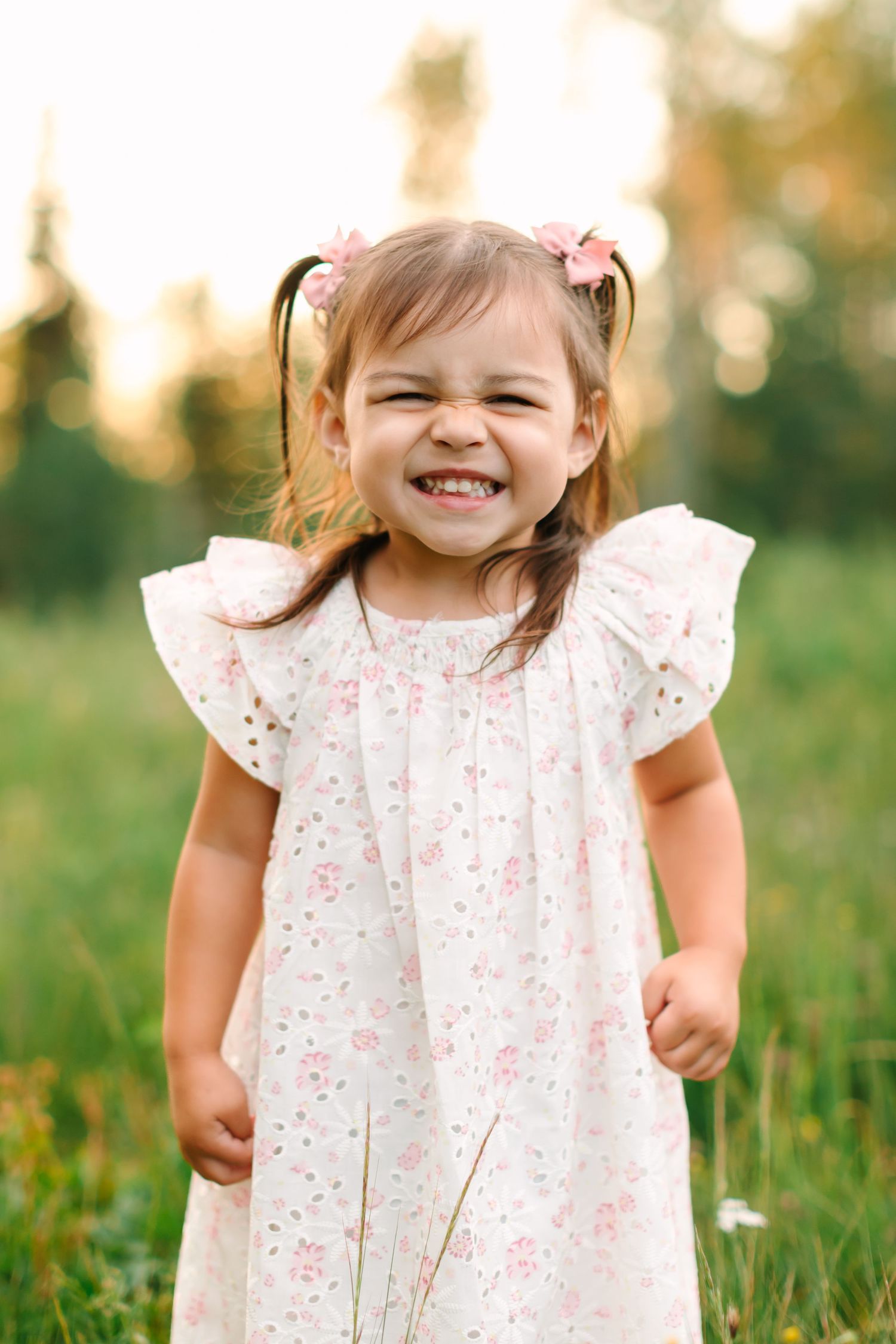 A cheerful toddler girl in a floral white dress with pink hair bows smiles broadly outdoors in a green field.