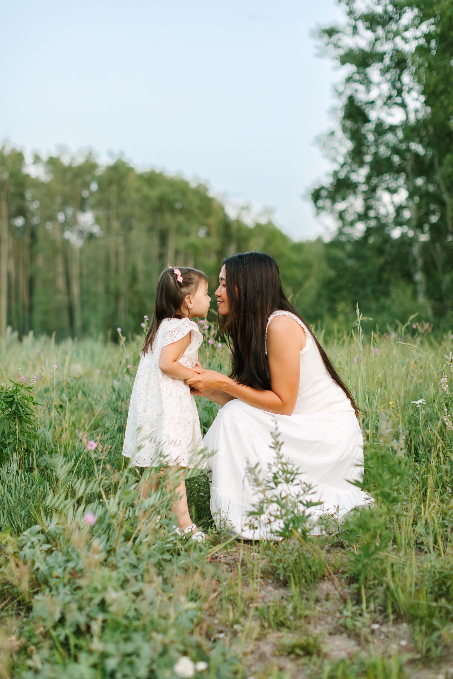 Mother and daughter in white dresses share a tender moment in a green meadow surrounded by wildflowers and trees.