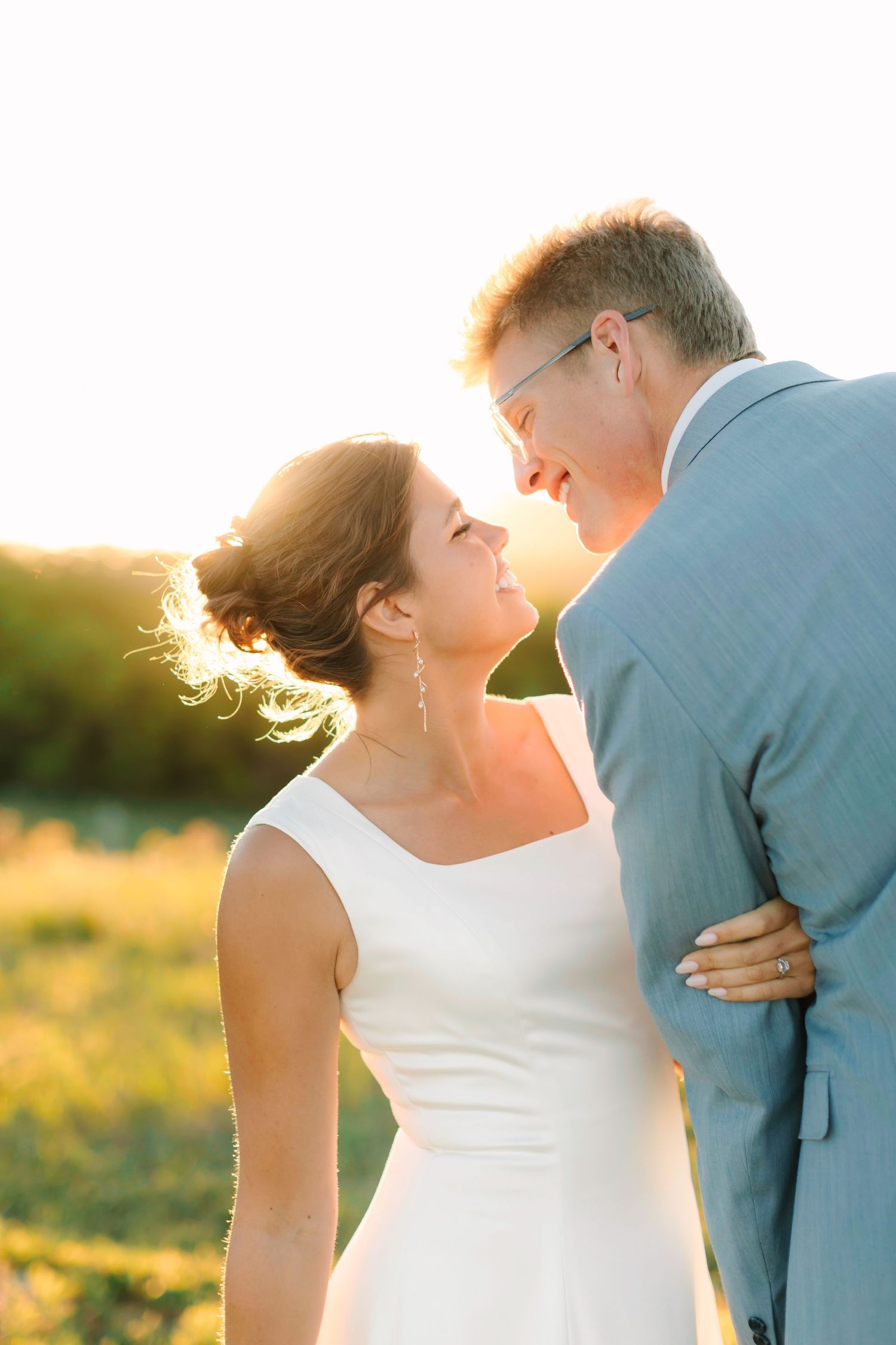 Bride and groom share romantic moment at sunset, smiling at each other in golden backlit outdoor setting.