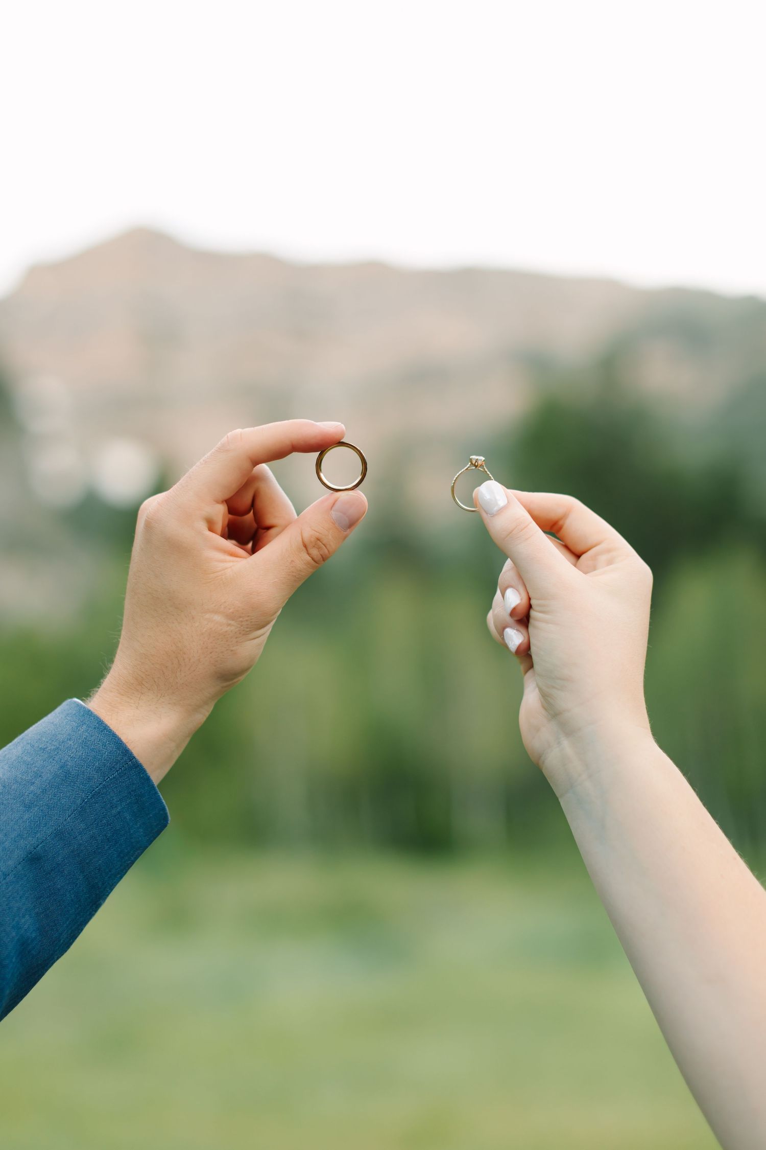 Bride and groom hold up their wedding rings outdoors with a blurred mountain landscape in the background.