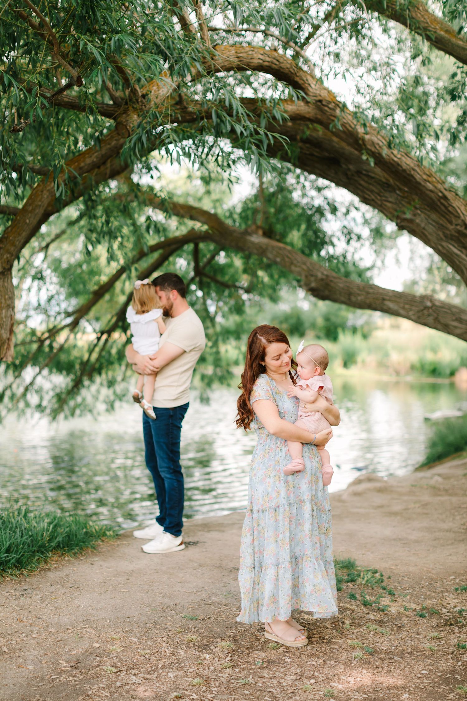 Family posing outdoors near a river under a willow tree, mother holding baby in blue dress, father with toddler.