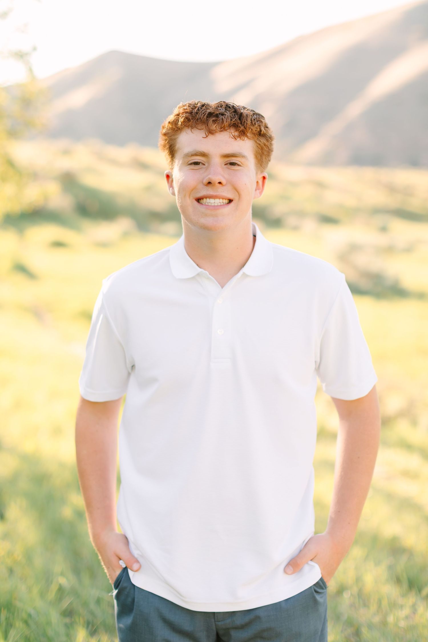 Smiling teenage boy with curly red hair wearing a white polo shirt standing in a sunny green field with mountains.