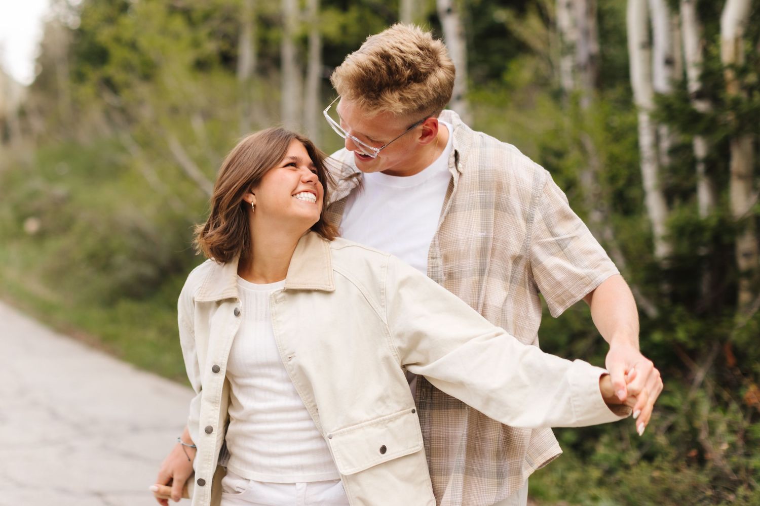 Happy young couple holding hands and smiling at each other while walking outdoors in a scenic wooded area.