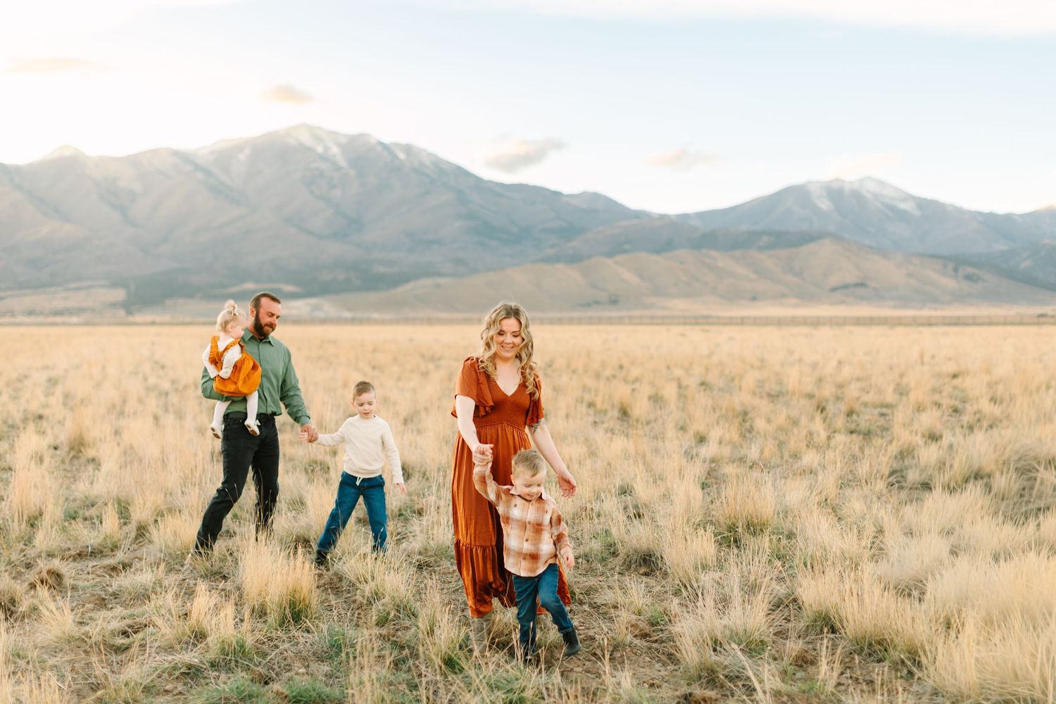 Couple embraces in golden field with mountains in background during sunset engagement photoshoot.