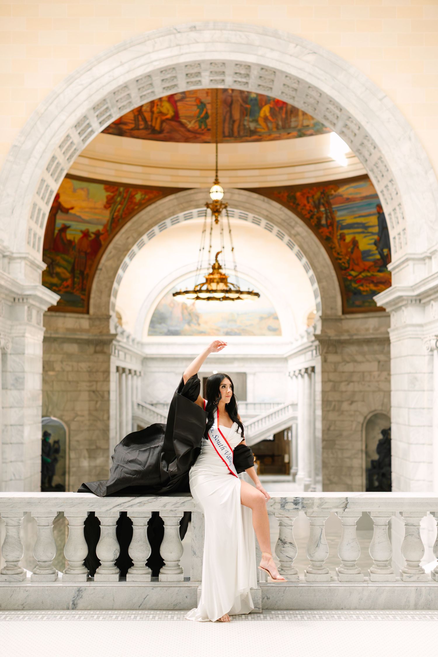 Romantic couple in white wedding attire poses beneath ornate arches with murals and classical architecture at Utah State Capitol.