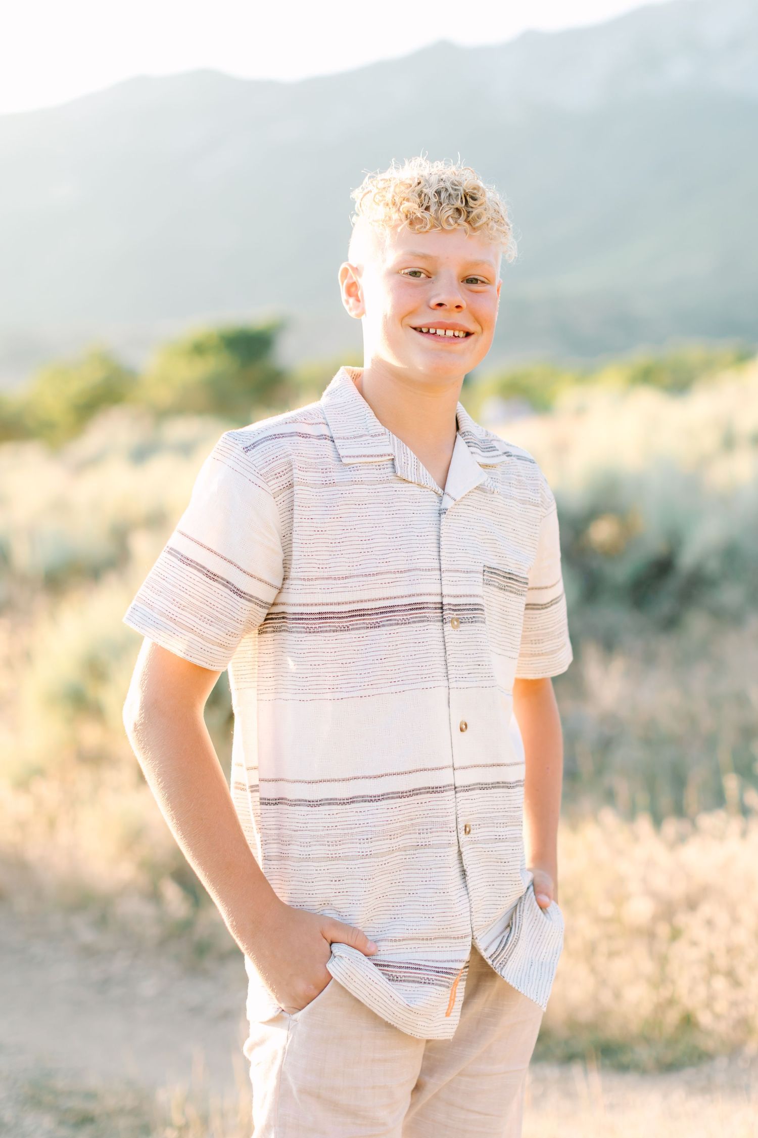 A child in a striped white shirt poses outdoors against a mountain backdrop in soft golden light.