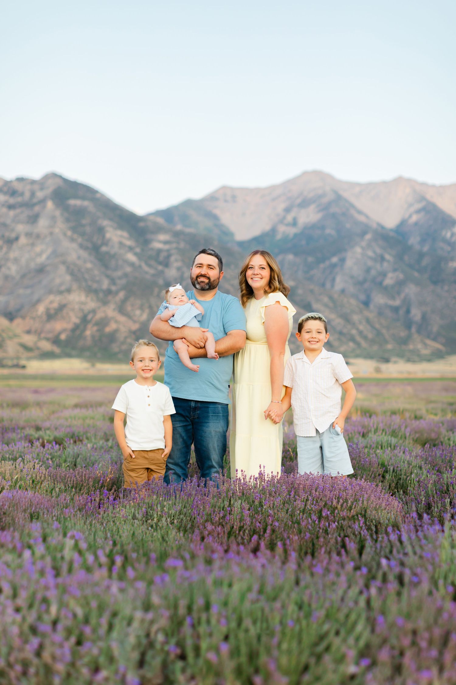 A family poses together in a blooming purple lavender field with majestic mountains in the background at sunset.