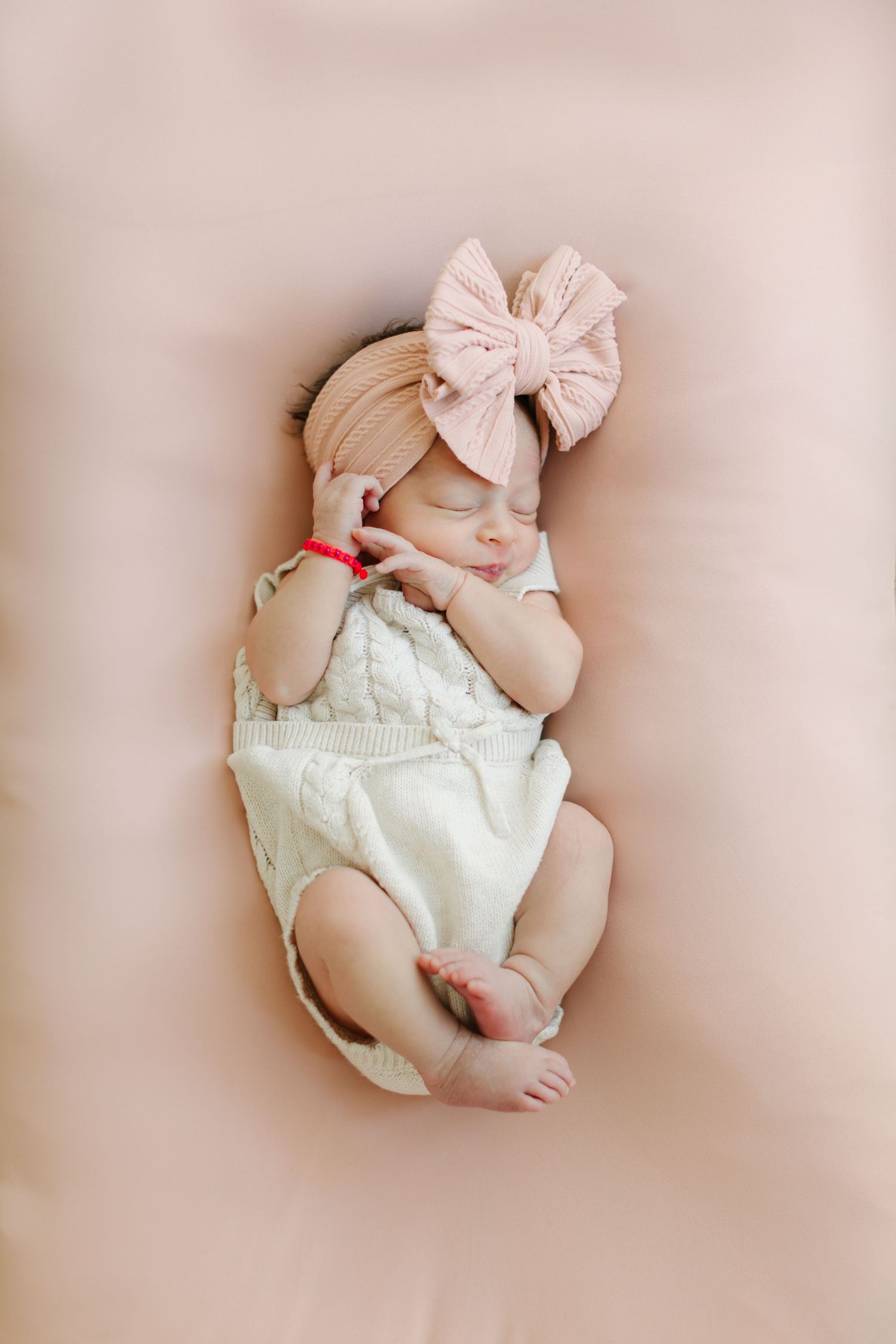 Sleeping newborn baby wearing white outfit and pink bow photographed on soft pink background.