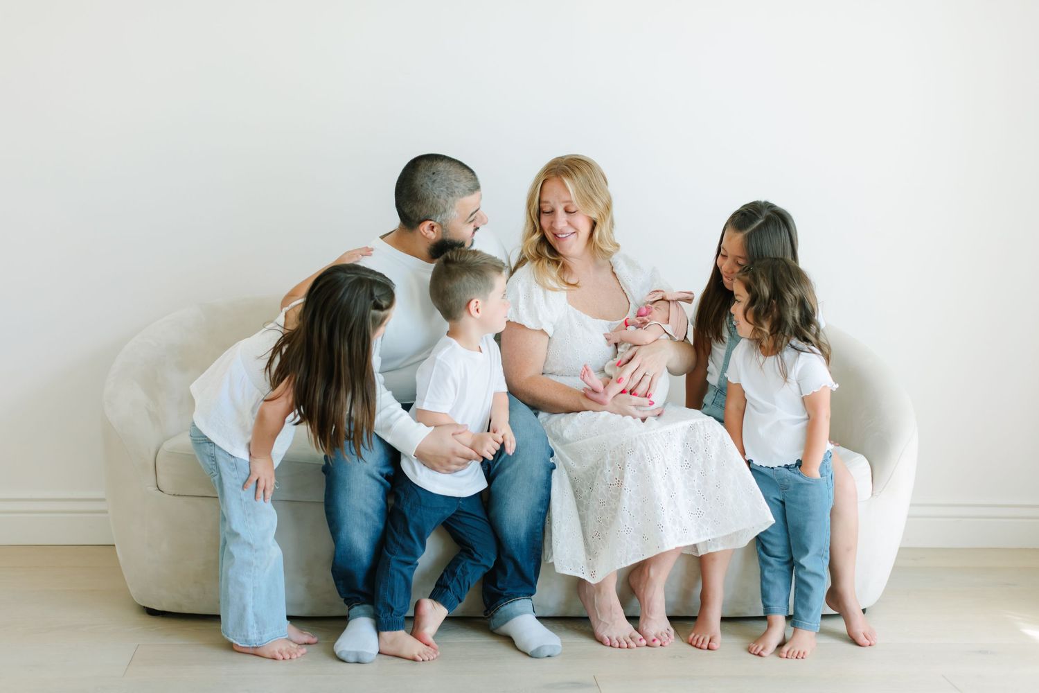 Family wearing coordinated white and denim outfits sits together on a white couch in a bright studio setting.