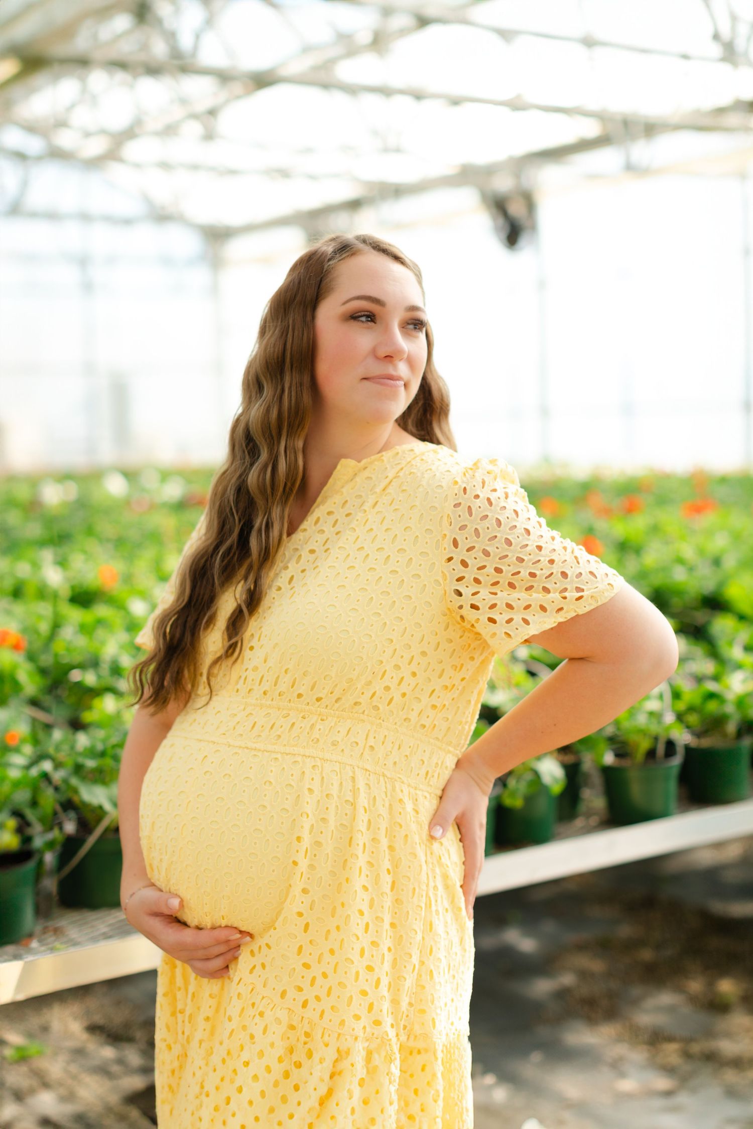 A woman in a yellow crochet dress poses in a sunny greenhouse surrounded by potted plants and flowers.