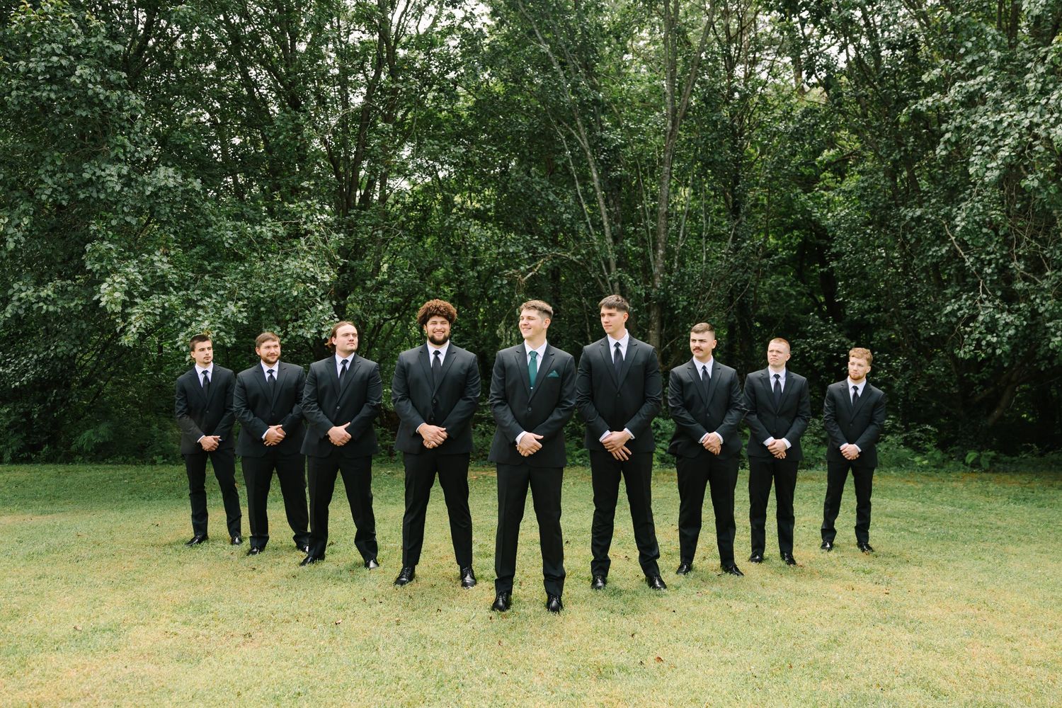 Wedding party members in black suits stand in a row on green grass surrounded by trees.