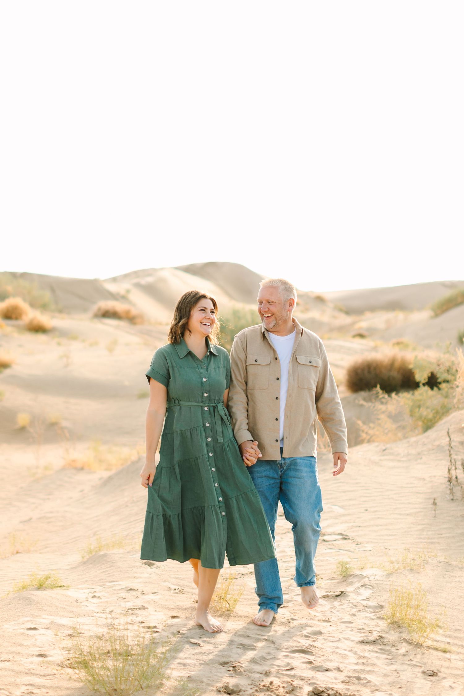 A couple walks hand in hand along sandy desert dunes during golden hour in matching casual outfits.