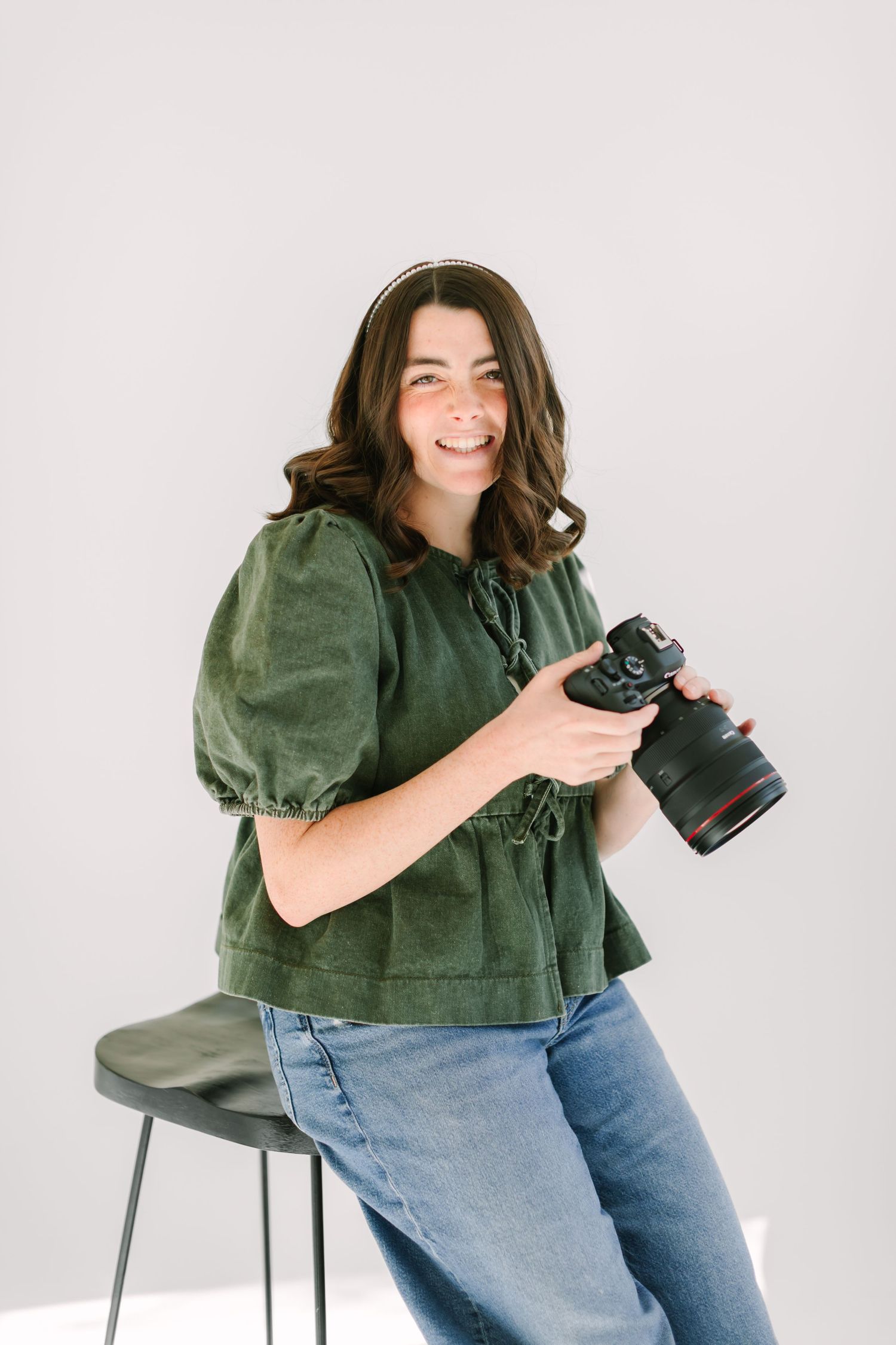 Photographer in green blouse and jeans sits on chair while reviewing photos on DSLR camera in studio setting.