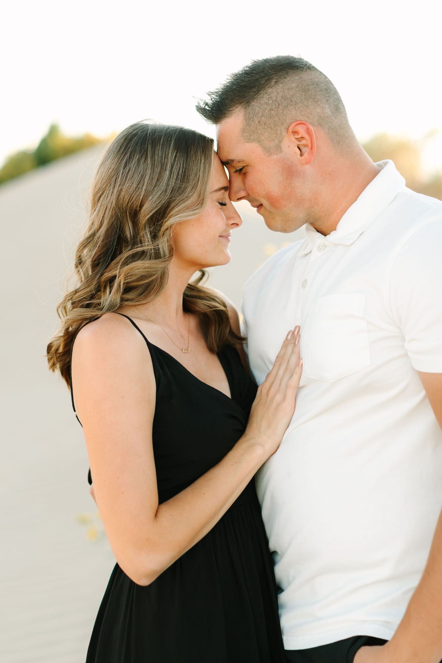 Romantic couple in black dress and white shirt embrace intimately during golden hour sunset.