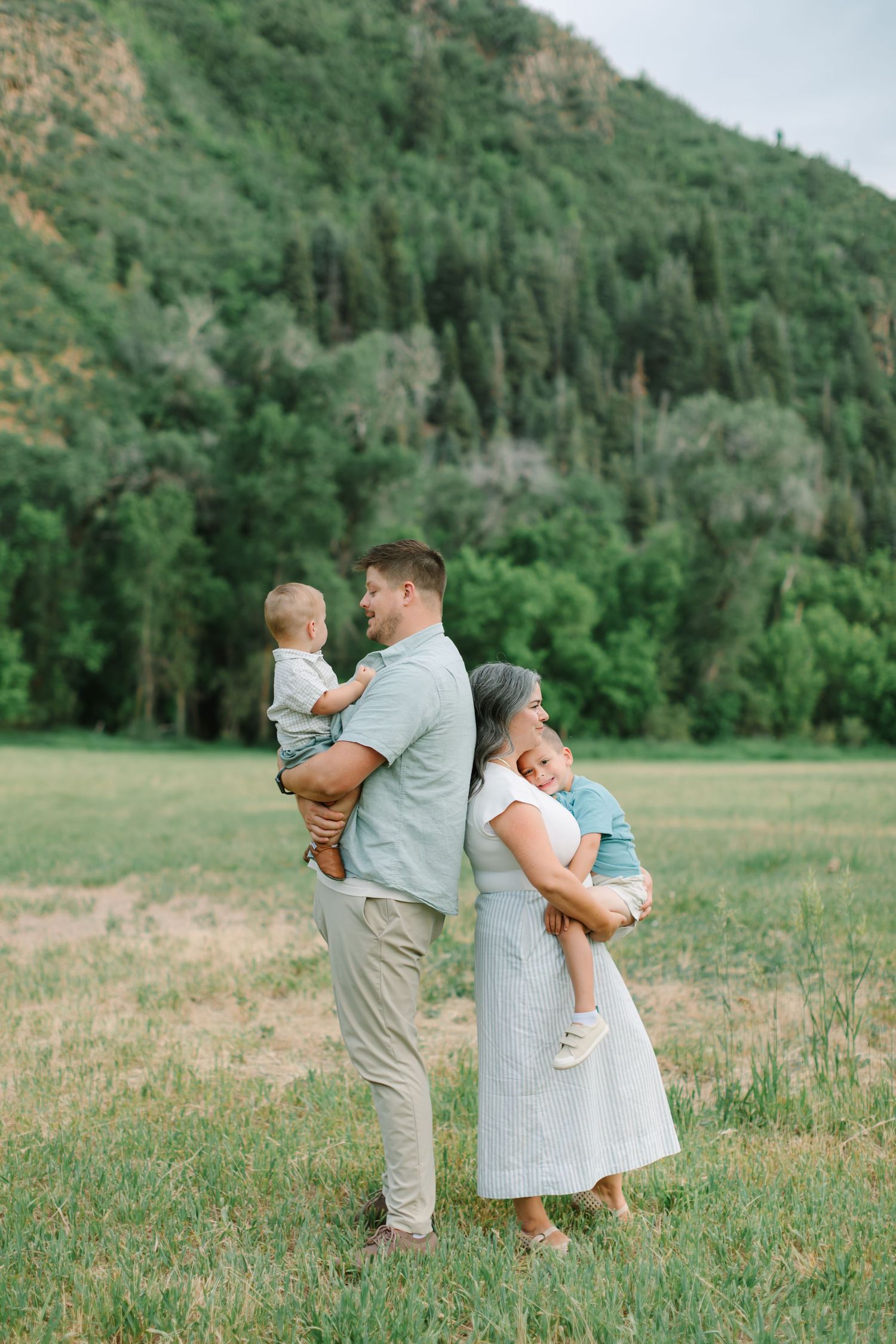 Family enjoying a tender moment together in a scenic mountain meadow surrounded by evergreen trees and rolling hills.
