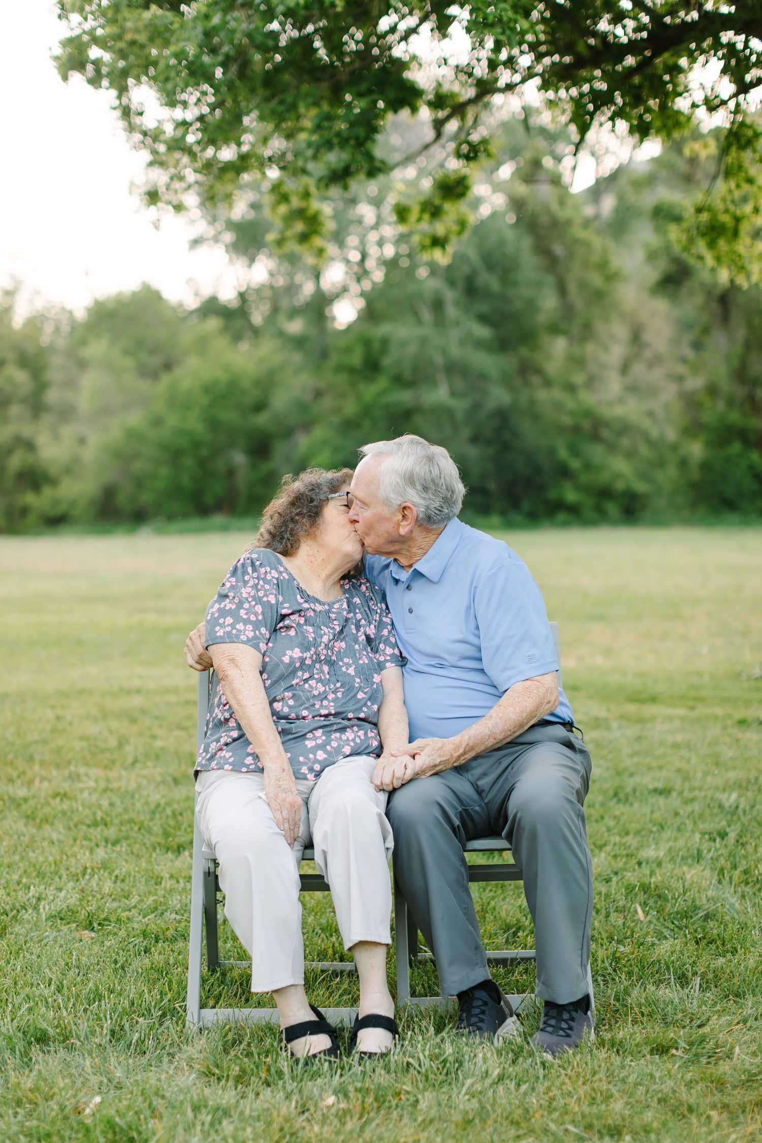 An elderly couple shares a tender moment while sitting together on a white bench in a sunny meadow.