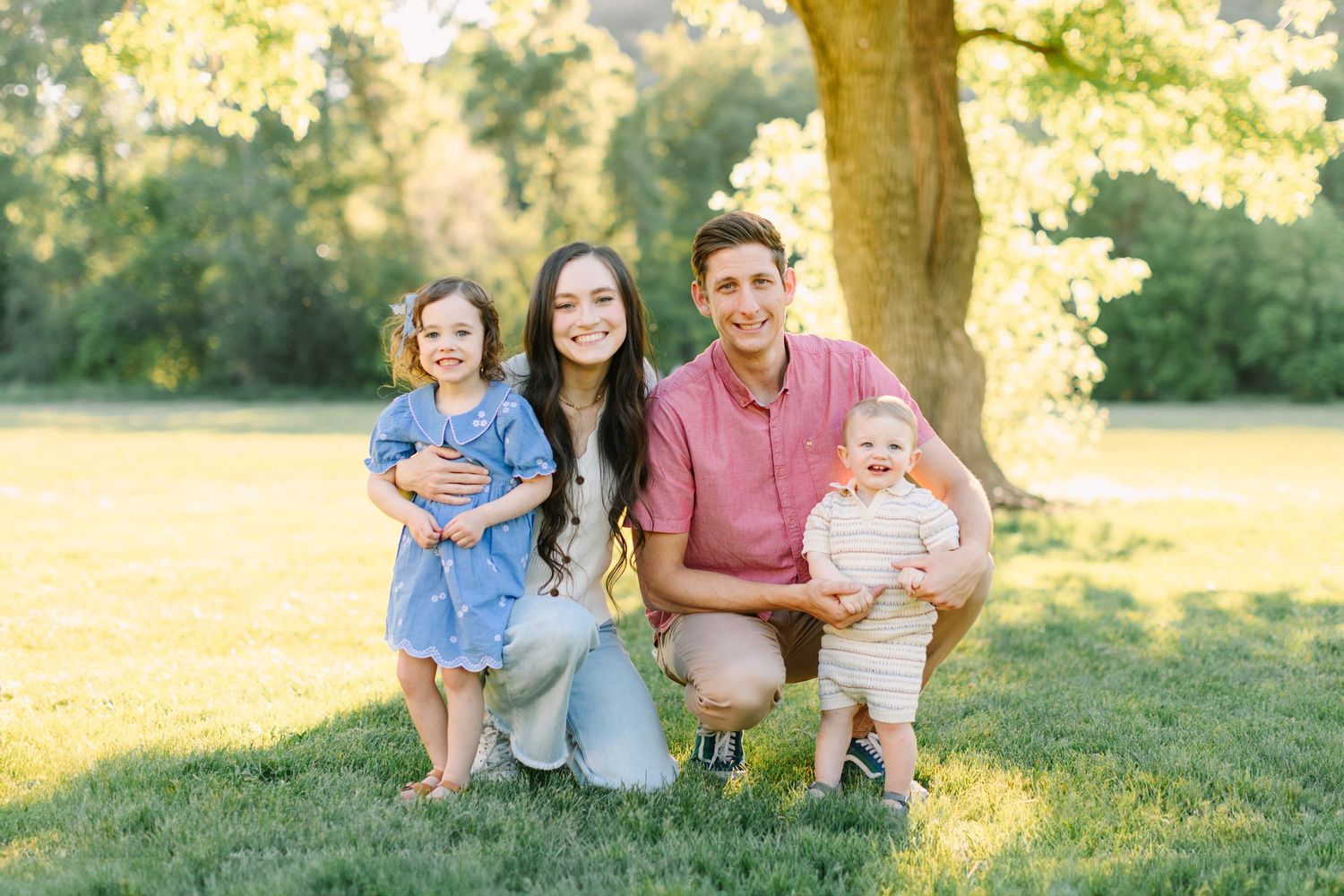 Family group sits together on grassy hill during golden hour sunset photo session in park.