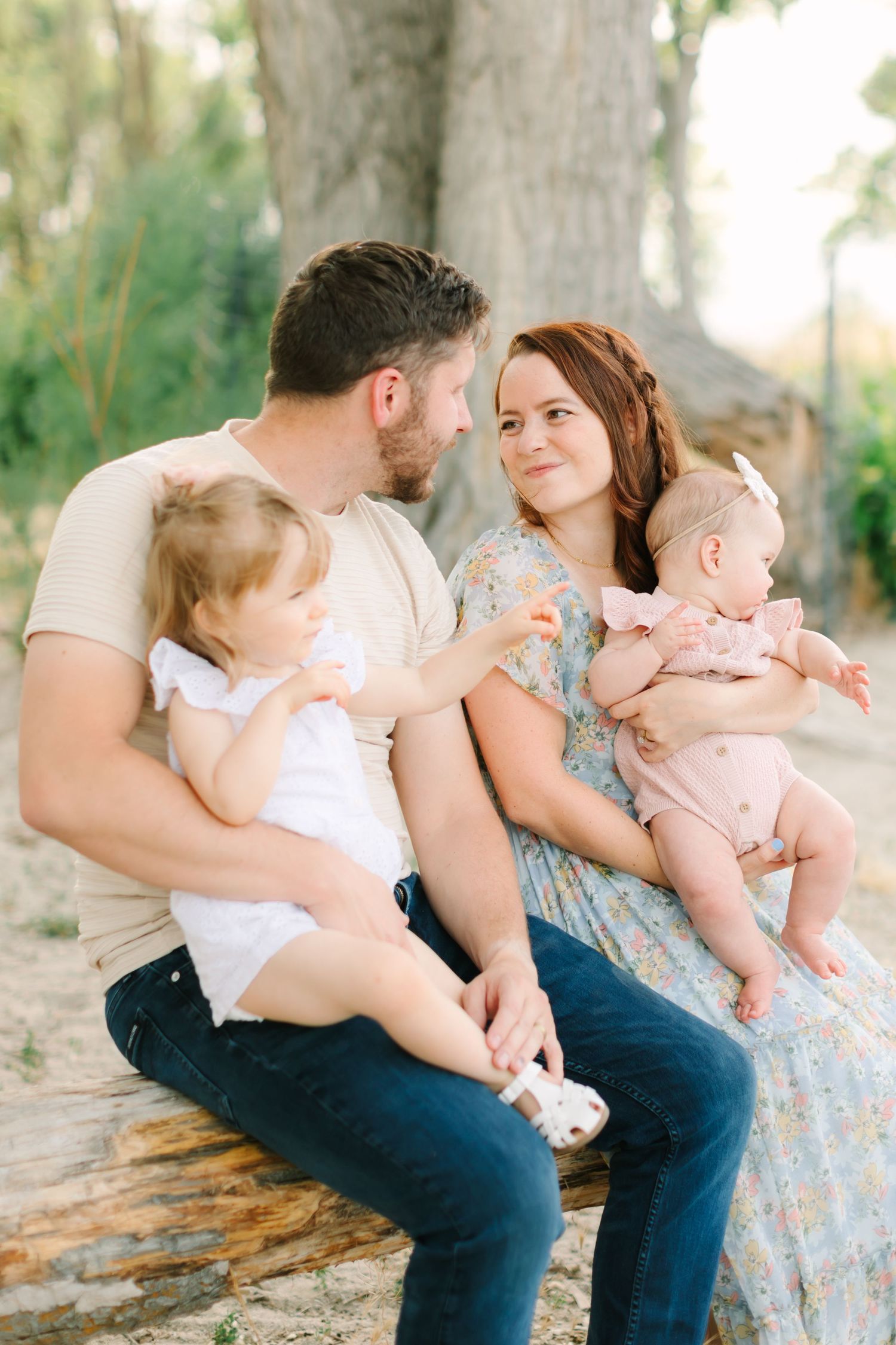 A young family of four sits on a log outdoors, parents gazing at each other while holding two small children.