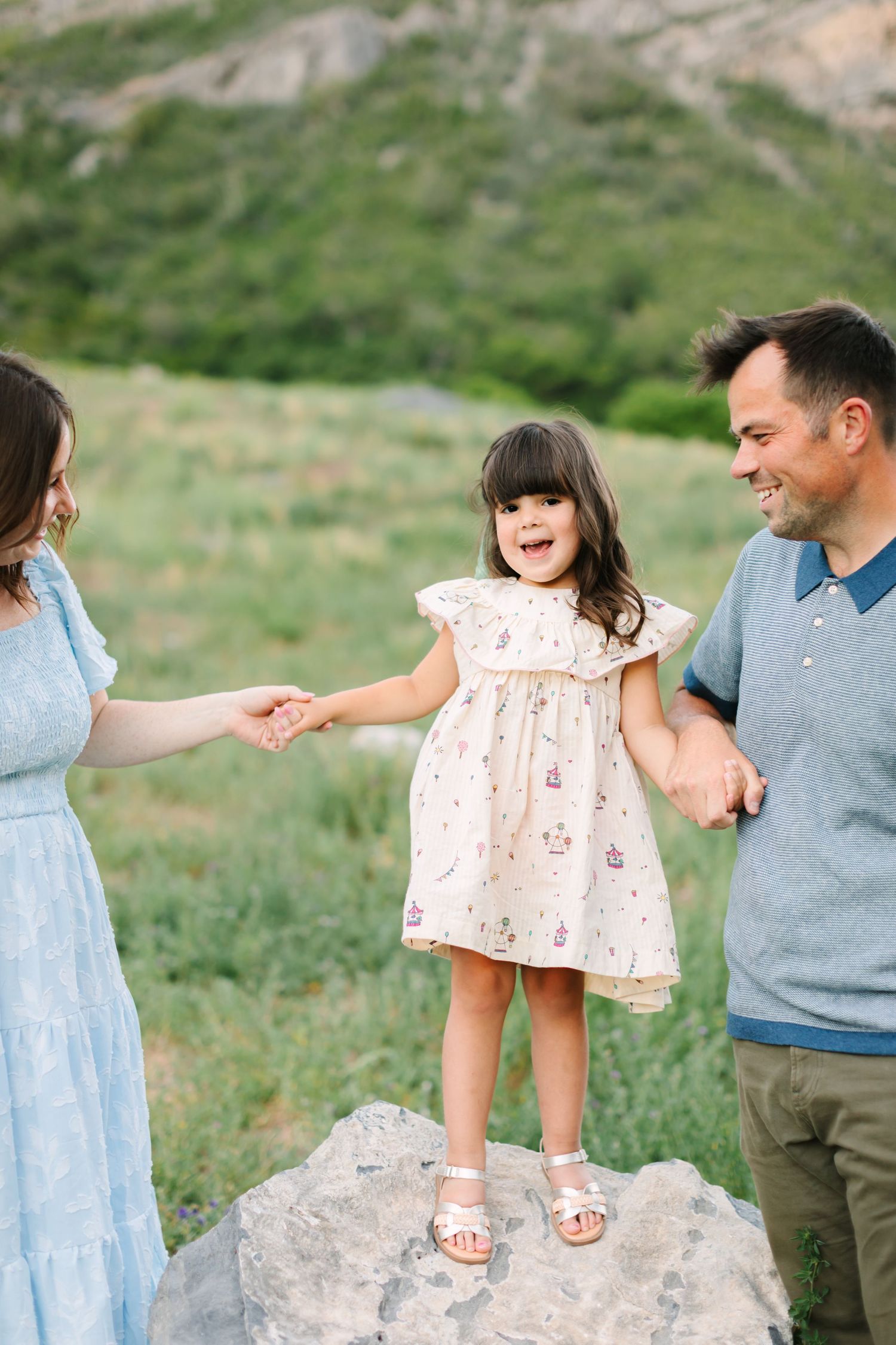 A joyful family of three holds hands outdoors, with a laughing little girl standing on a rock between her smiling parents.